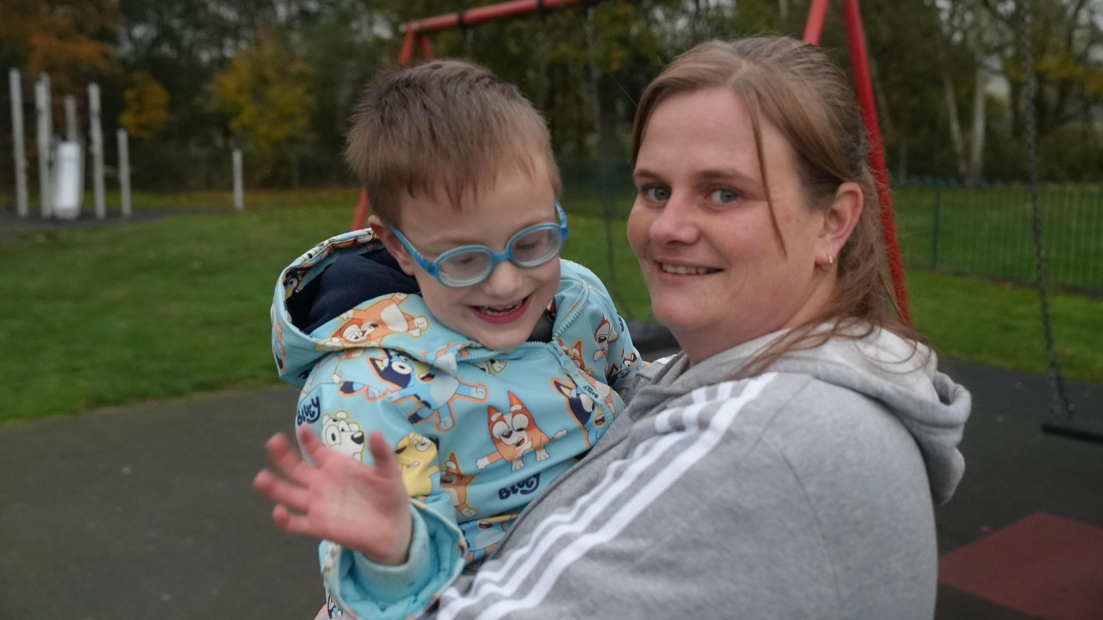 A woman is holding a boy in her arms. They are both smiling at the camera and stand in front of some swings 