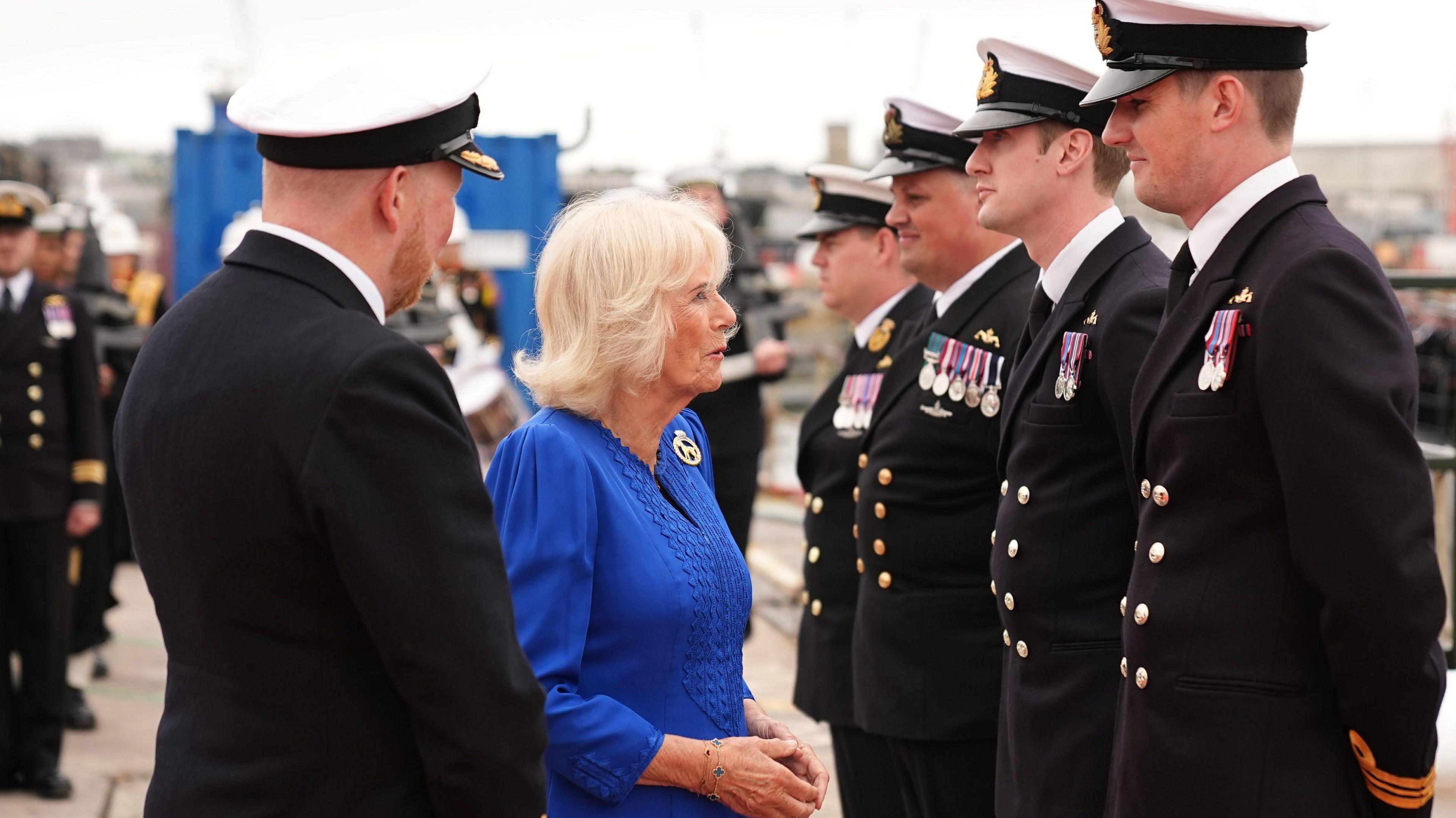 Queen Camilla, a woman with shoulder-length grey hair in a blue dress, stands in front of a line of submariners in dress uniform, wearing white naval caps. An officer stands next to the Queen as she talks to one of the men.
