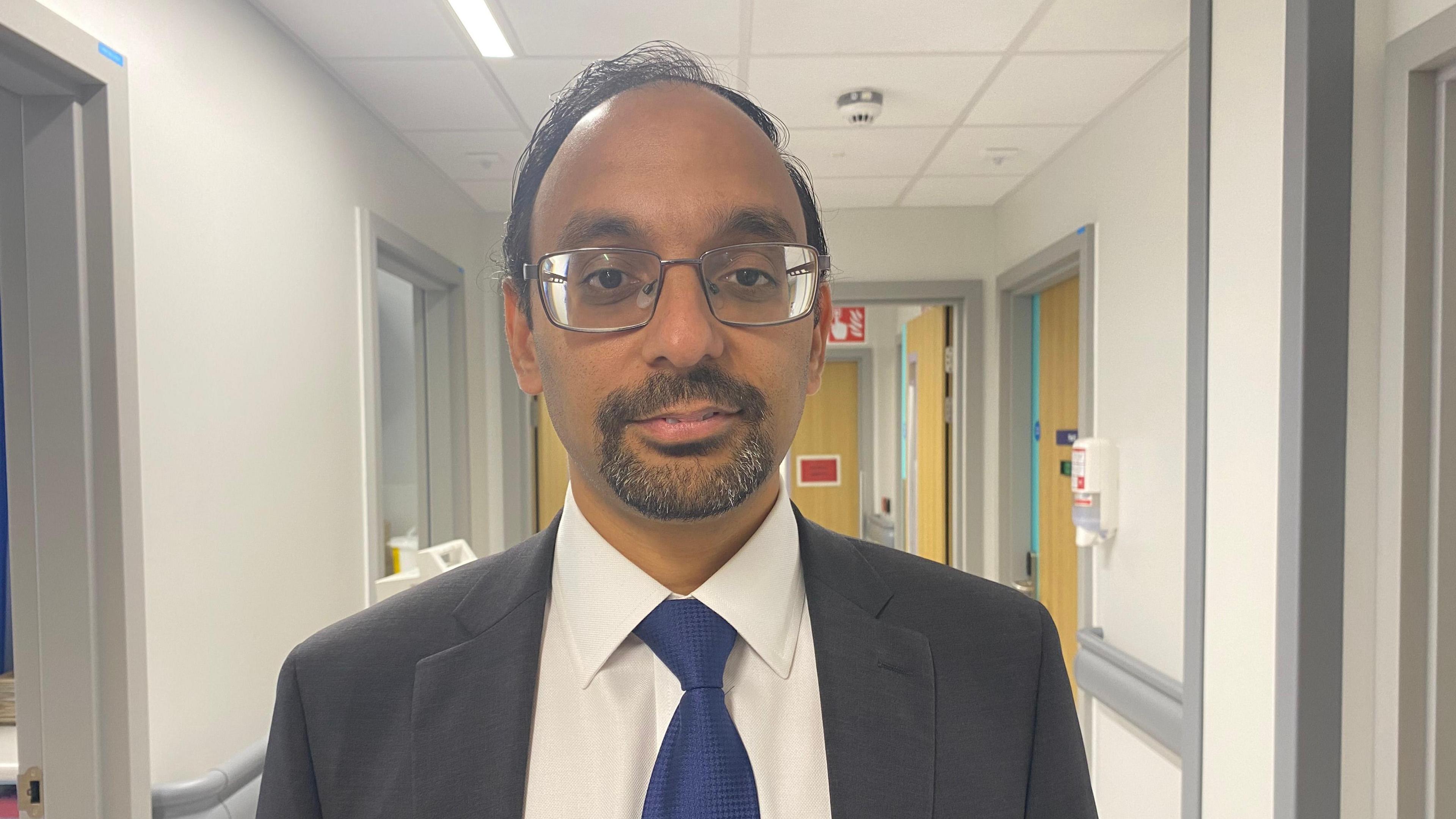 Saurav looking into the camera and smiling slightly. He's wearing a dark grey suit, with a white shirt and a dark blue tie. He's stood in a hospital corridor which has white walls and grey doorframes.