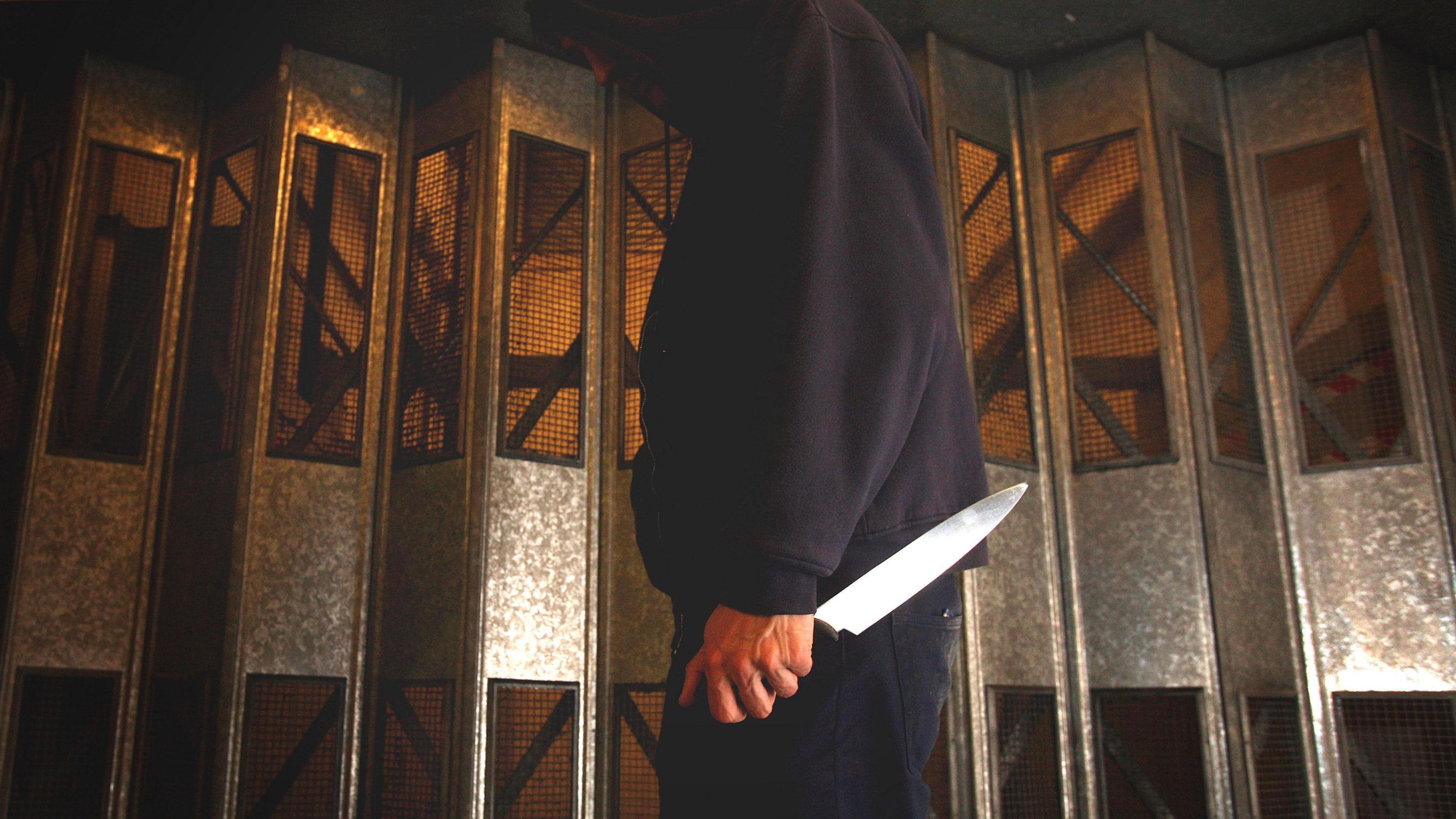A man in a dark blue hoody stands side on to the camera holding a large kitchen knife in his left hand.