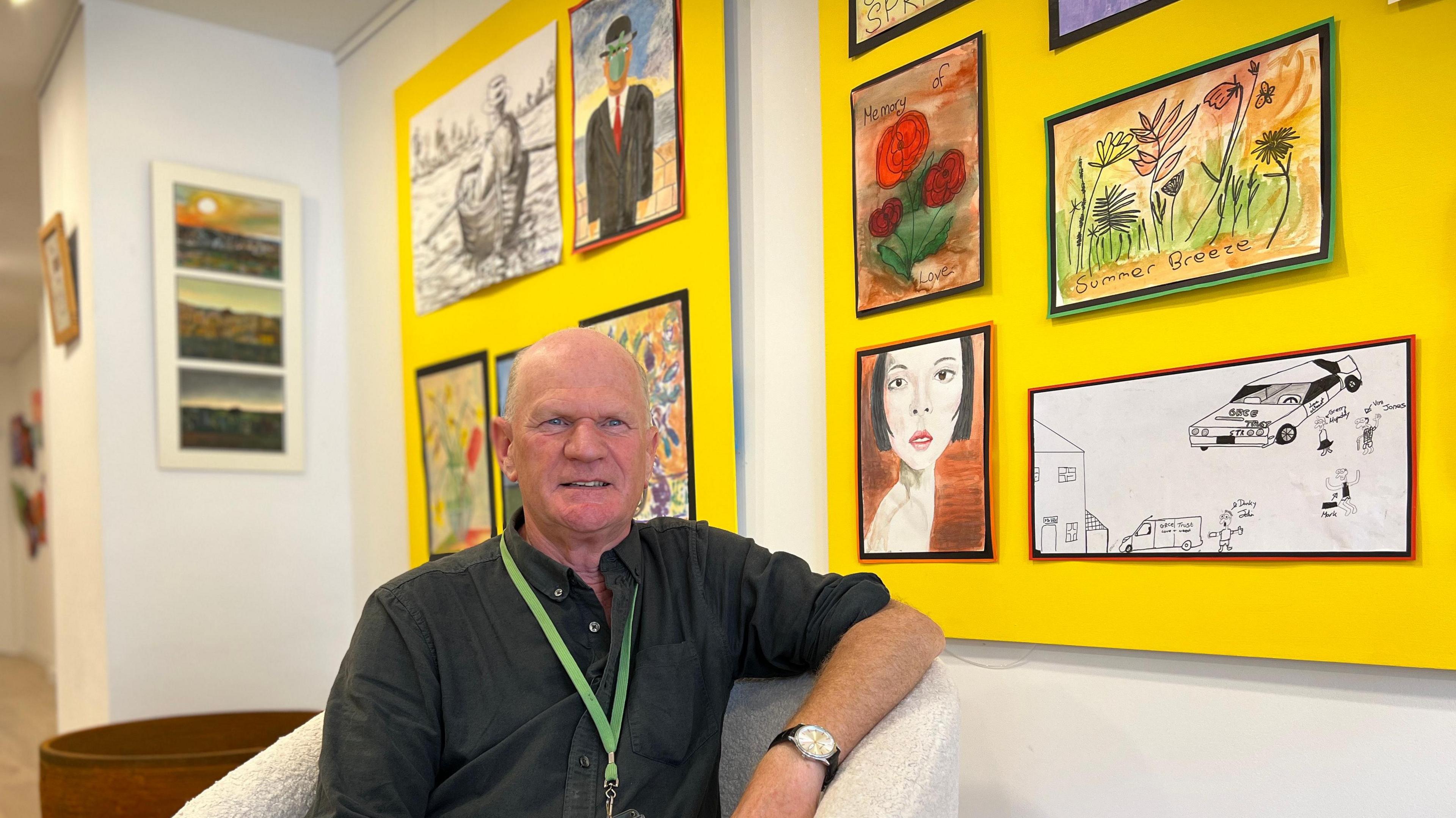 A bald man sits on a chair with artwork behind him.