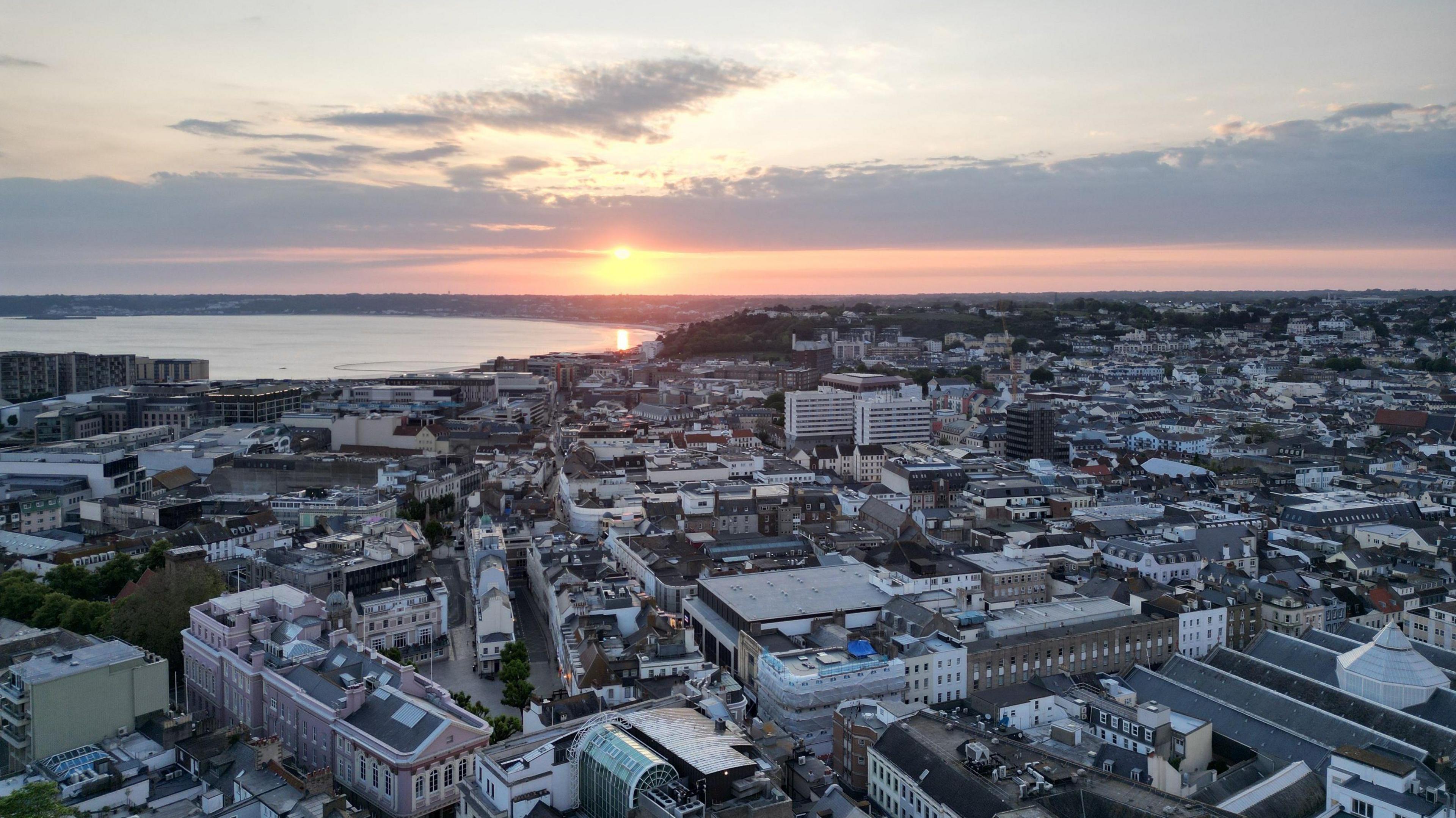 Aerial view of a cityscape at sunset. The sun is setting over the horizon, casting a warm glow across the sky and reflecting on the water in the distance. The city below is densely packed with buildings of various sizes and styles, with some greenery visible among them. The scene captures a serene and picturesque moment as day transitions to night
