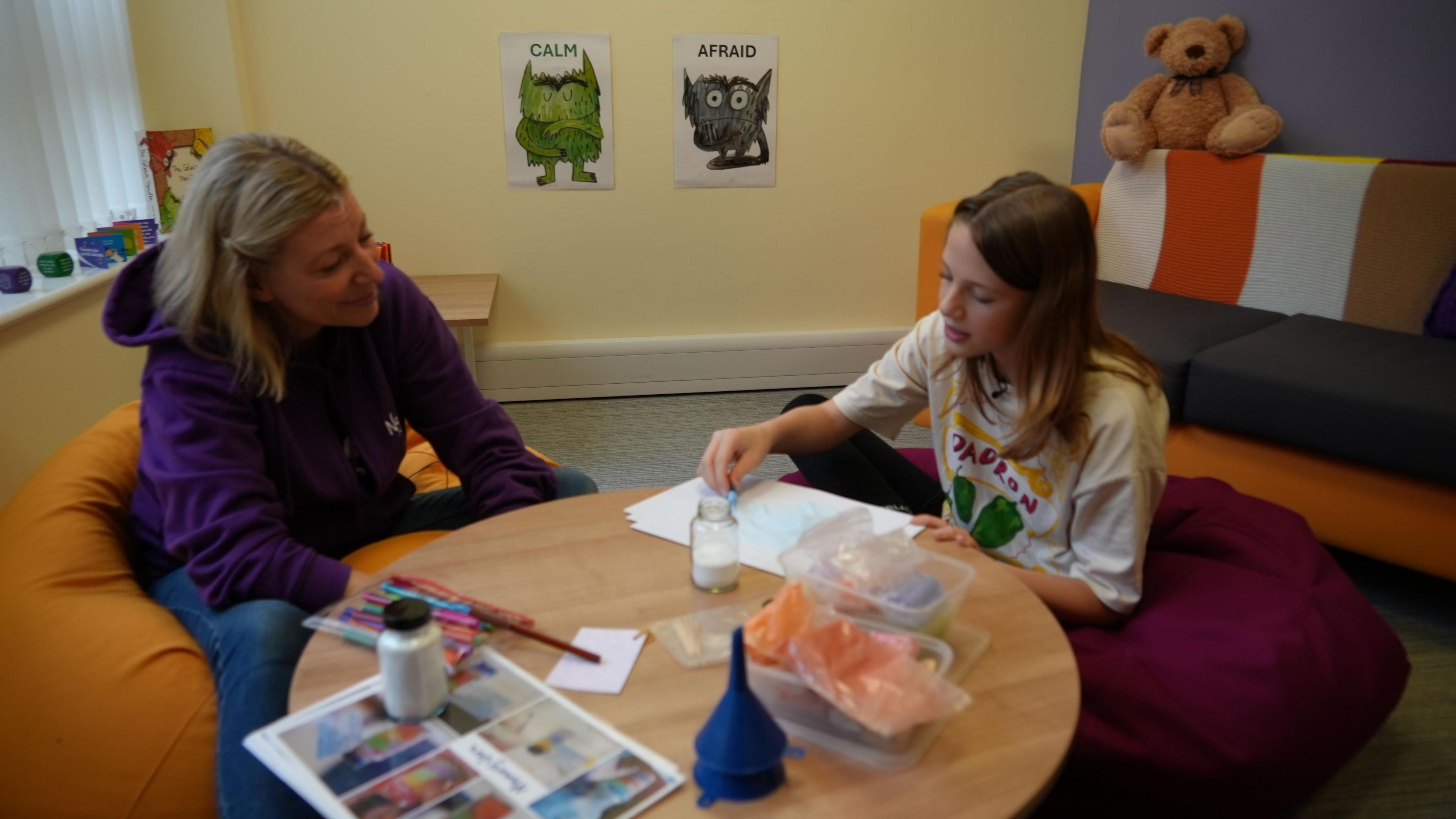 Woman wearing purple hoodie seated by a craft table next to a teenage girl wearing a white patterned t-shirt.  They are making memory jars using coloured sand.