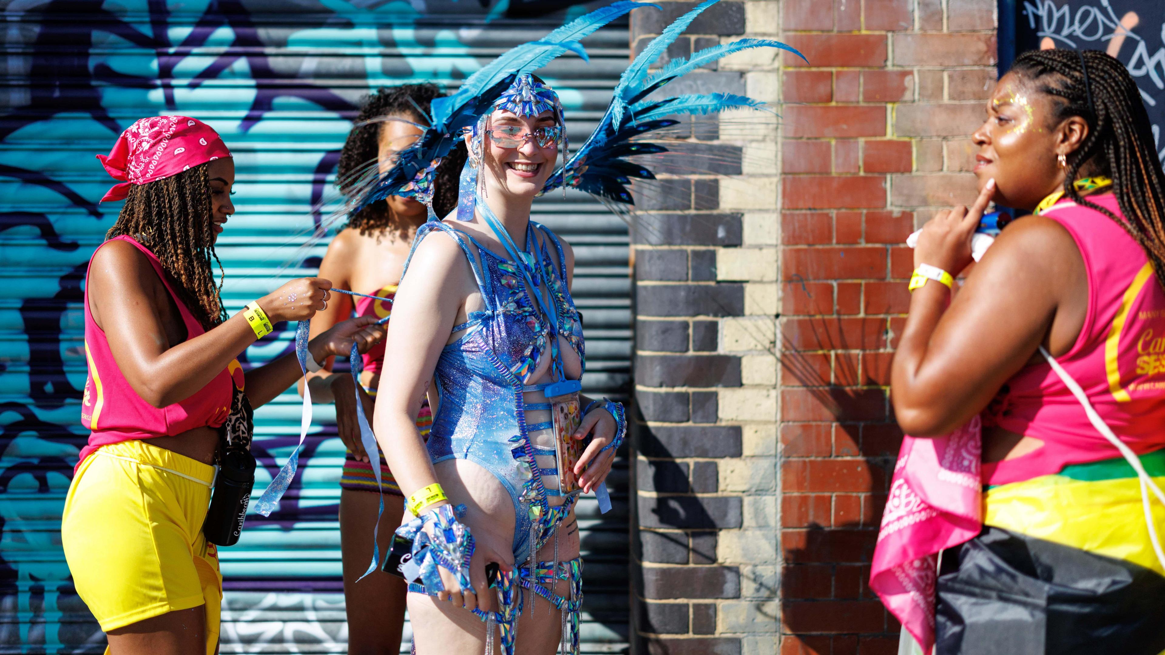 A woman in a blue costume is helped with the ties of her outfit by two other women wearing pink and yellow. 