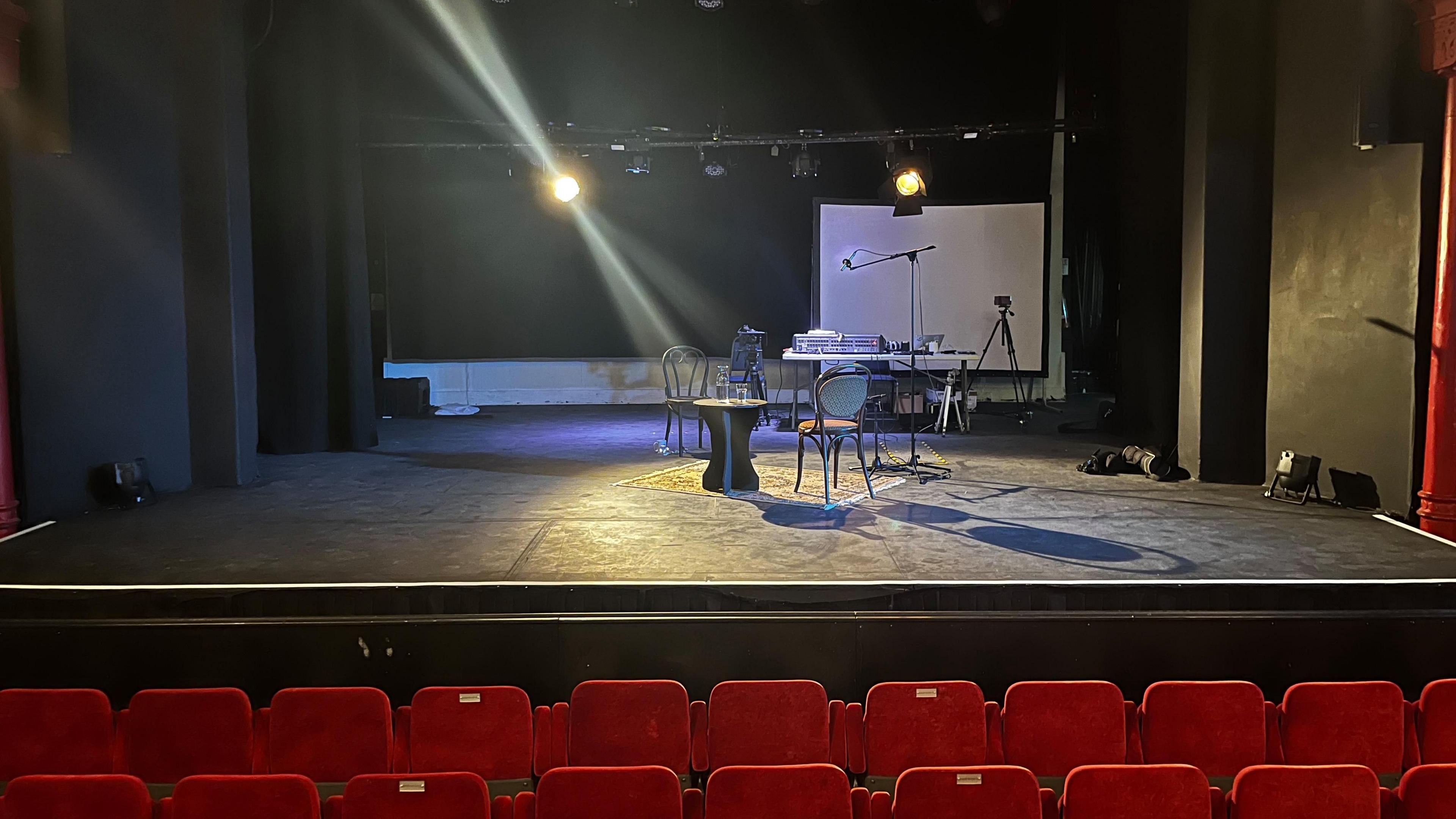The inside of a theatre, showing the stage. There are red seats at the front. On the stage is a table and chairs. Several lights shine into shot.