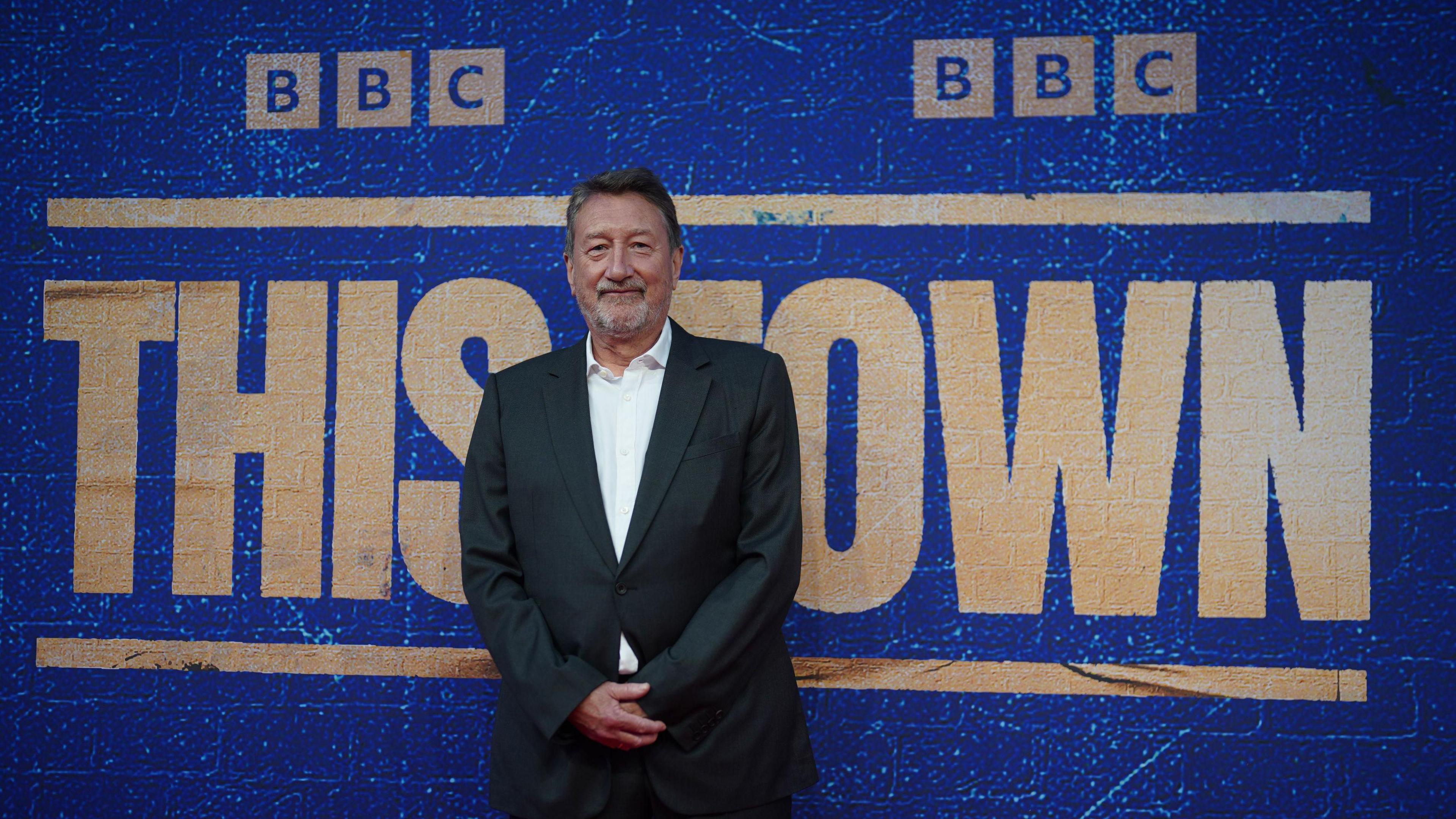 Steven Knight is wearing a black suit and is standing in front of a large blue backdrop. On the backdrop in big letters it reads "BBC This Town"