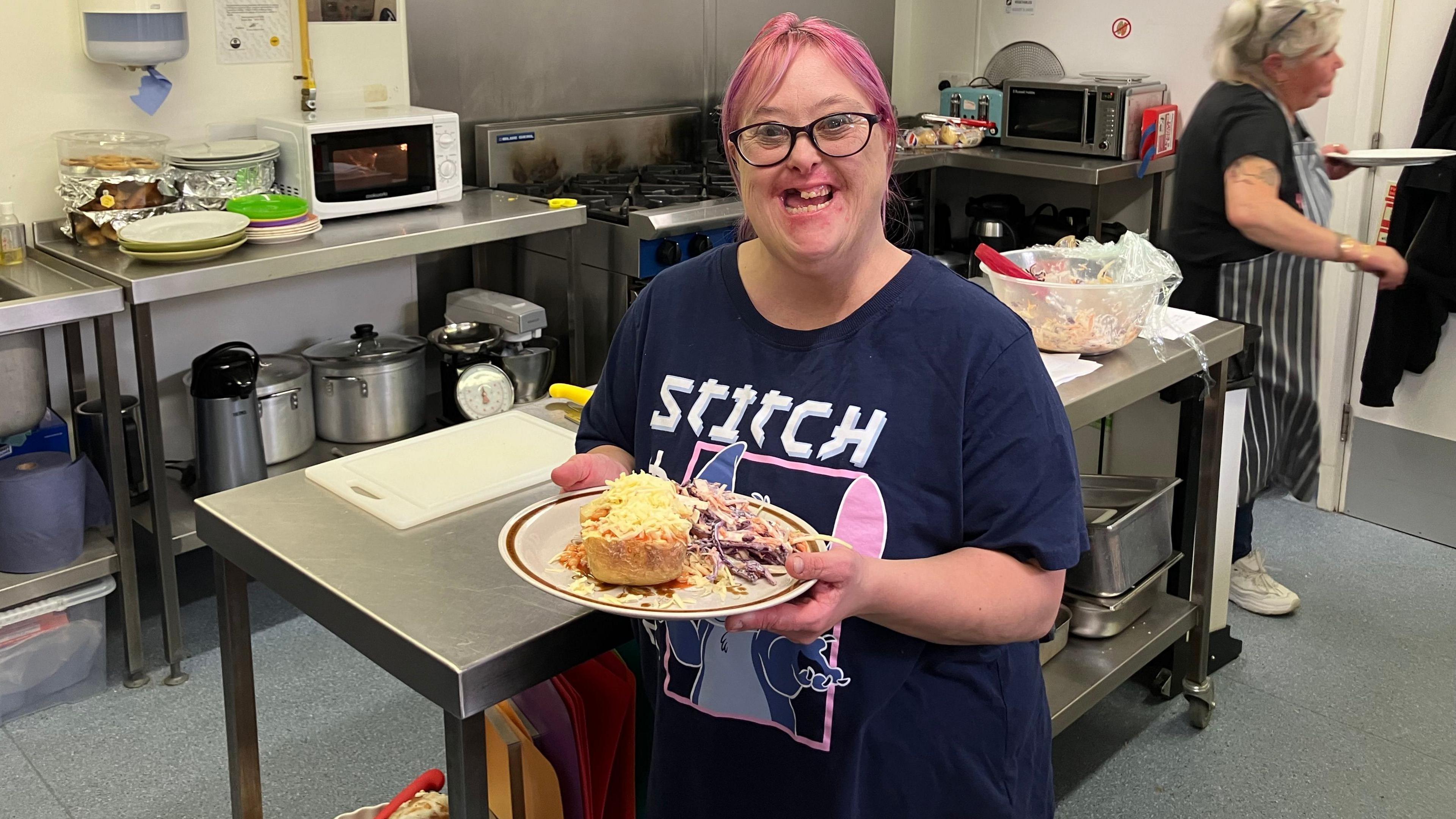 Liz,  a volunteer at the cafe- wearing a blue T-shirt- pink hair- holding a plate of jacket potato