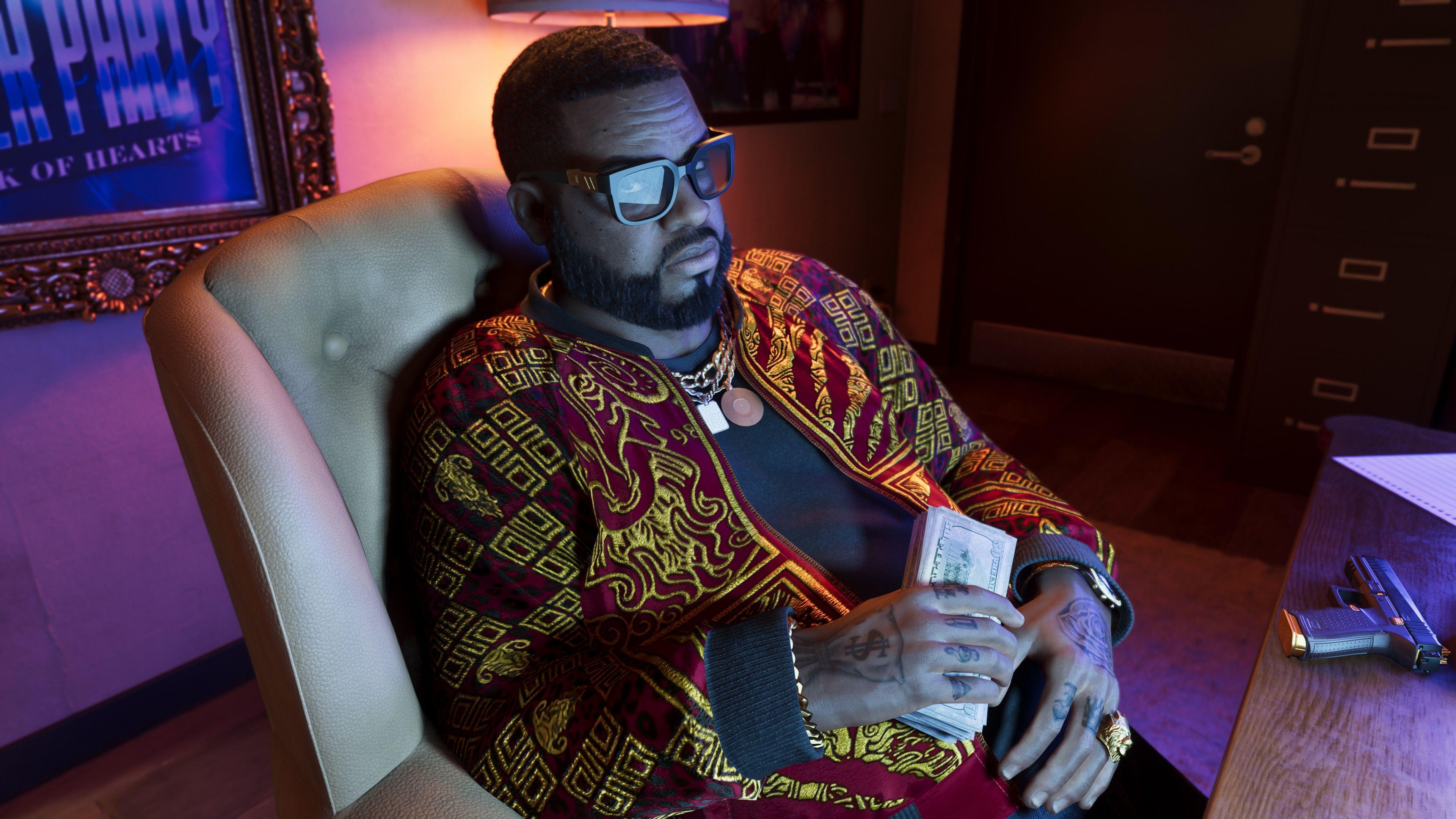 A man sits in a dimly lit office, holding a thick wad of banknotes in his hand. He wears a burgundy jacket with elaborate gold patterning, gold chains and a chunky ring on his finger. The light of a computer monitor reflects off his black-rimmed glasses and a pistol lies on the desk in front of him.