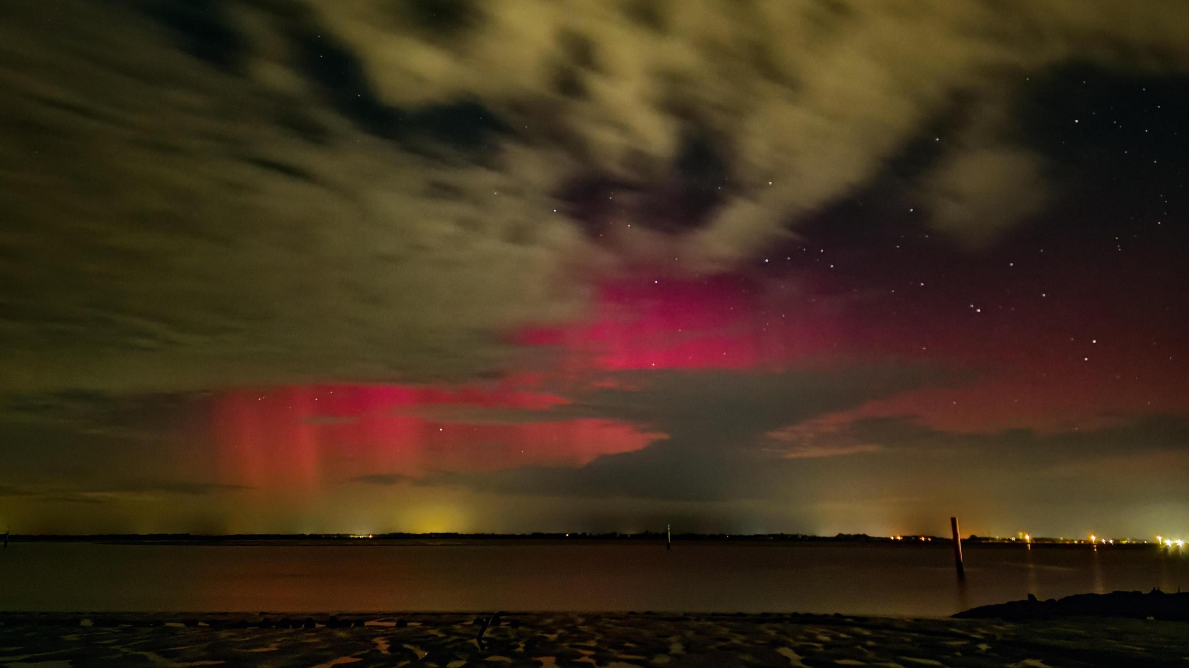 image shows the aurora borealis as a deep pink scad on the sky and the shoreline below