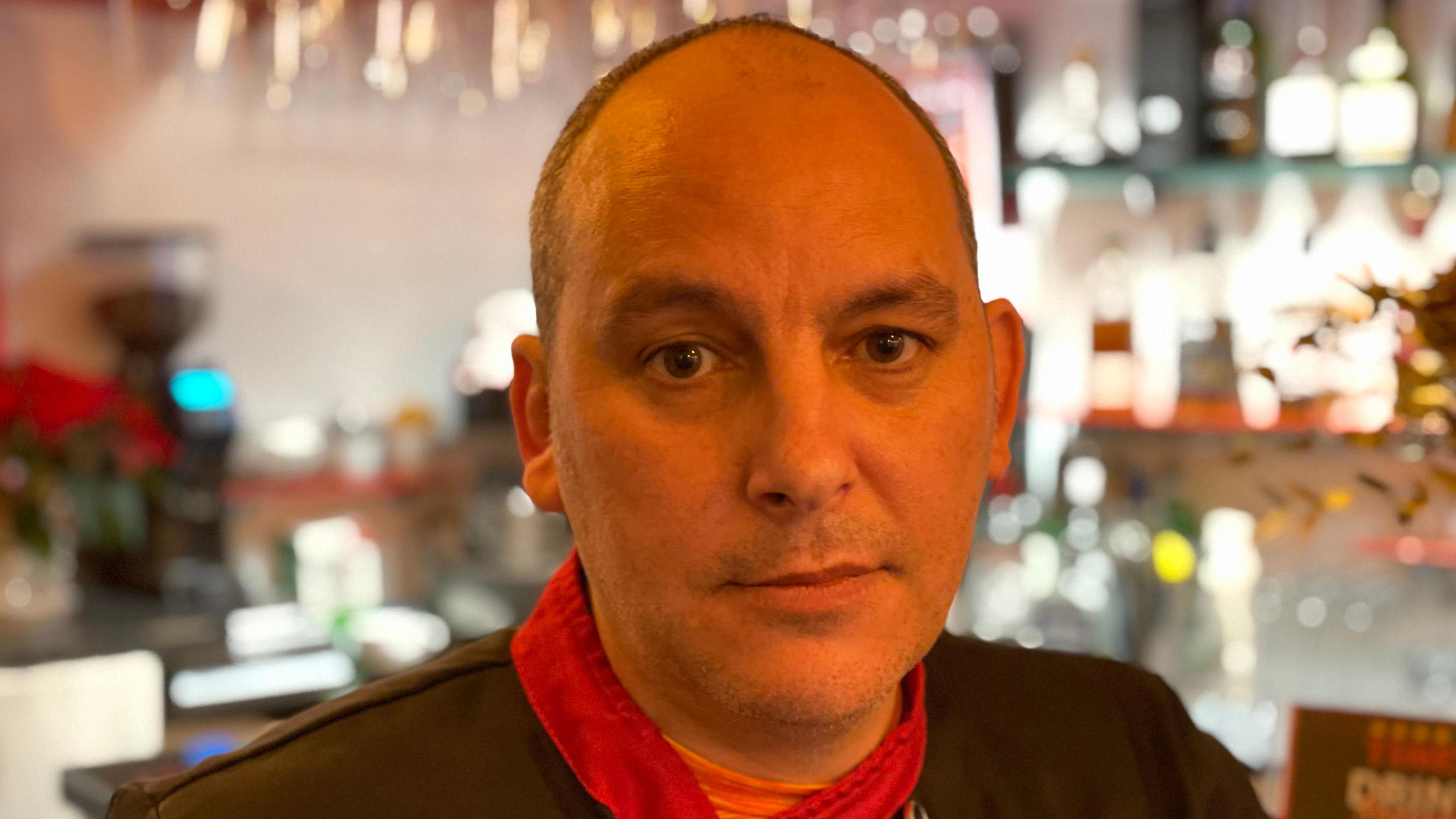 Flavio Serra looks into the camera. He has dark eyes and balding hair. He is wearing a dark chef's outfit with red trims. He is standing at the bar in his restaurant and bottles are visible in the background.