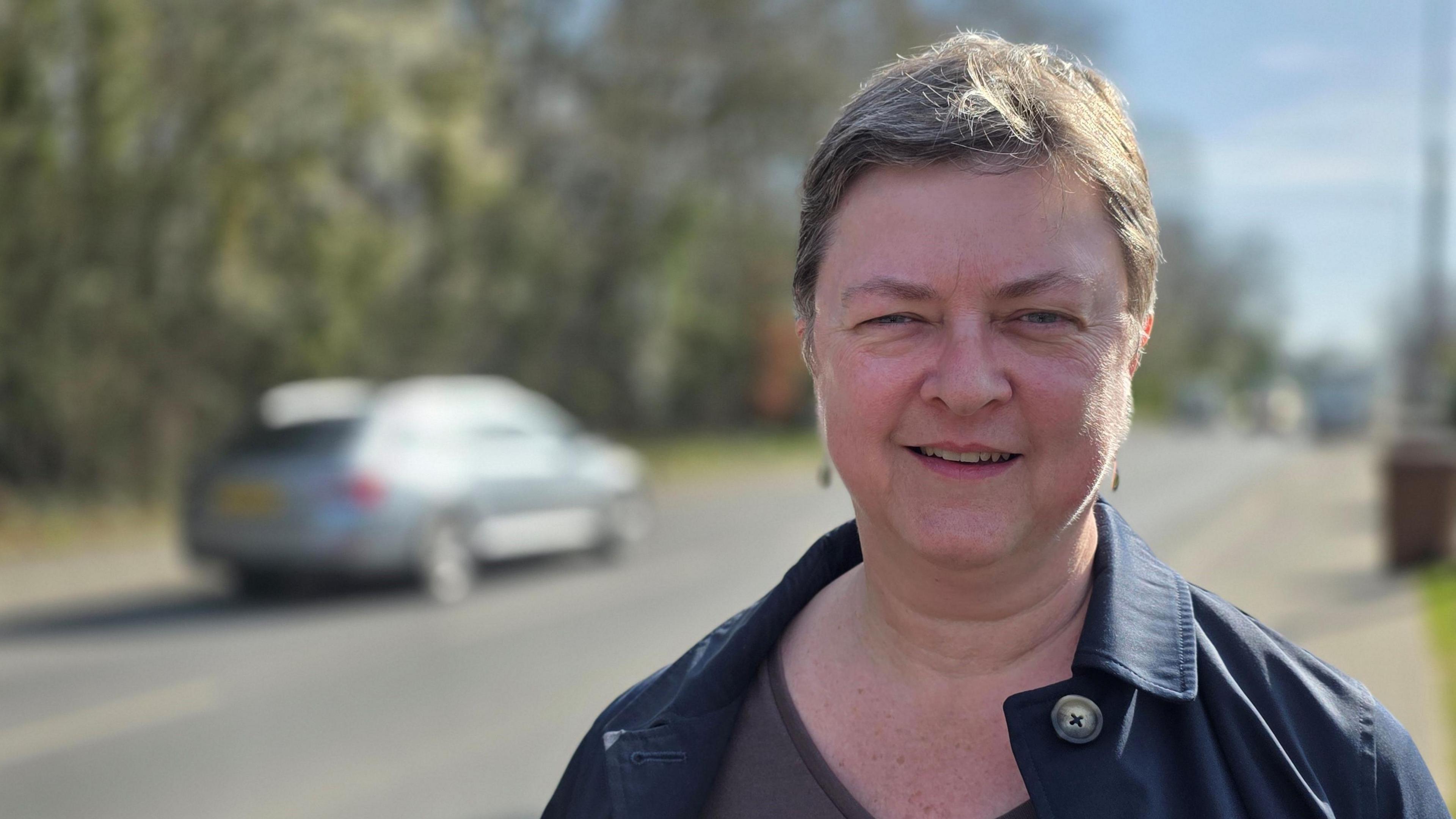 Nicola Fowler is smiling at the camera. She's wearing a blue coat. Behind her, we can see a car driving past.