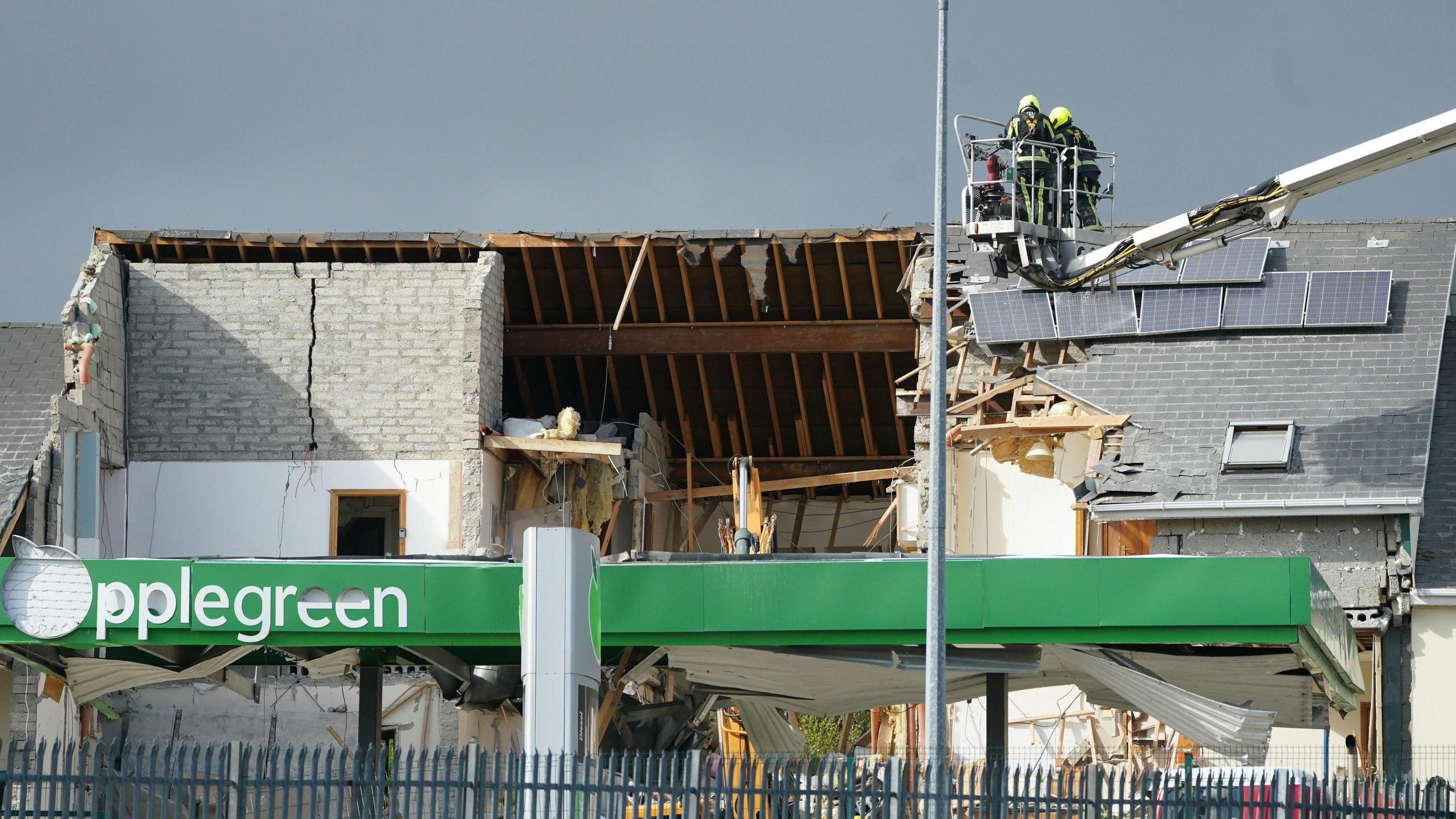 Two firefighters in an aerial platform appliance above the scene of the explosion at a service station in Creeslough in October 2022.  Most of the pitched roof of the front of the building has been blown off.  The inside of the wooden rafters are exposed, tiles are missing, solar panels have been displaced and there is a large crack in a grey brick wall which reaches the pitch of the roof. 
