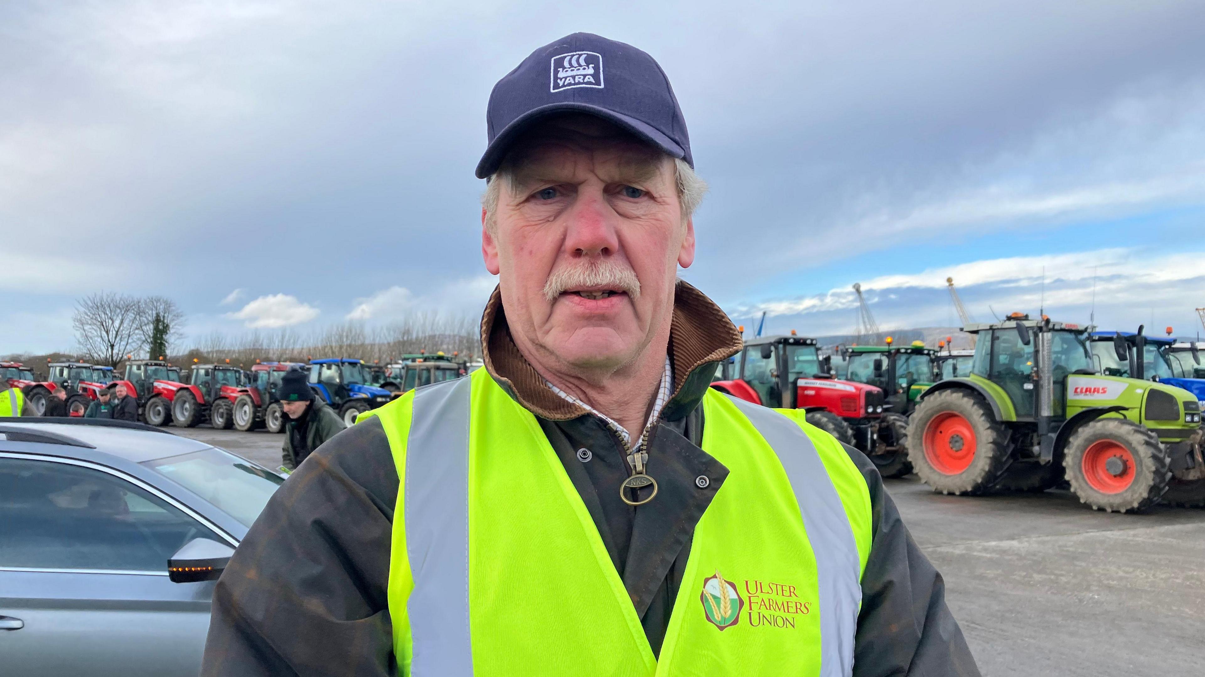 A man, with grey hair and a grey moustache, wearing a jacket and hi-vis vest. He is also wearing a navy cap. There are tractors in the background. 