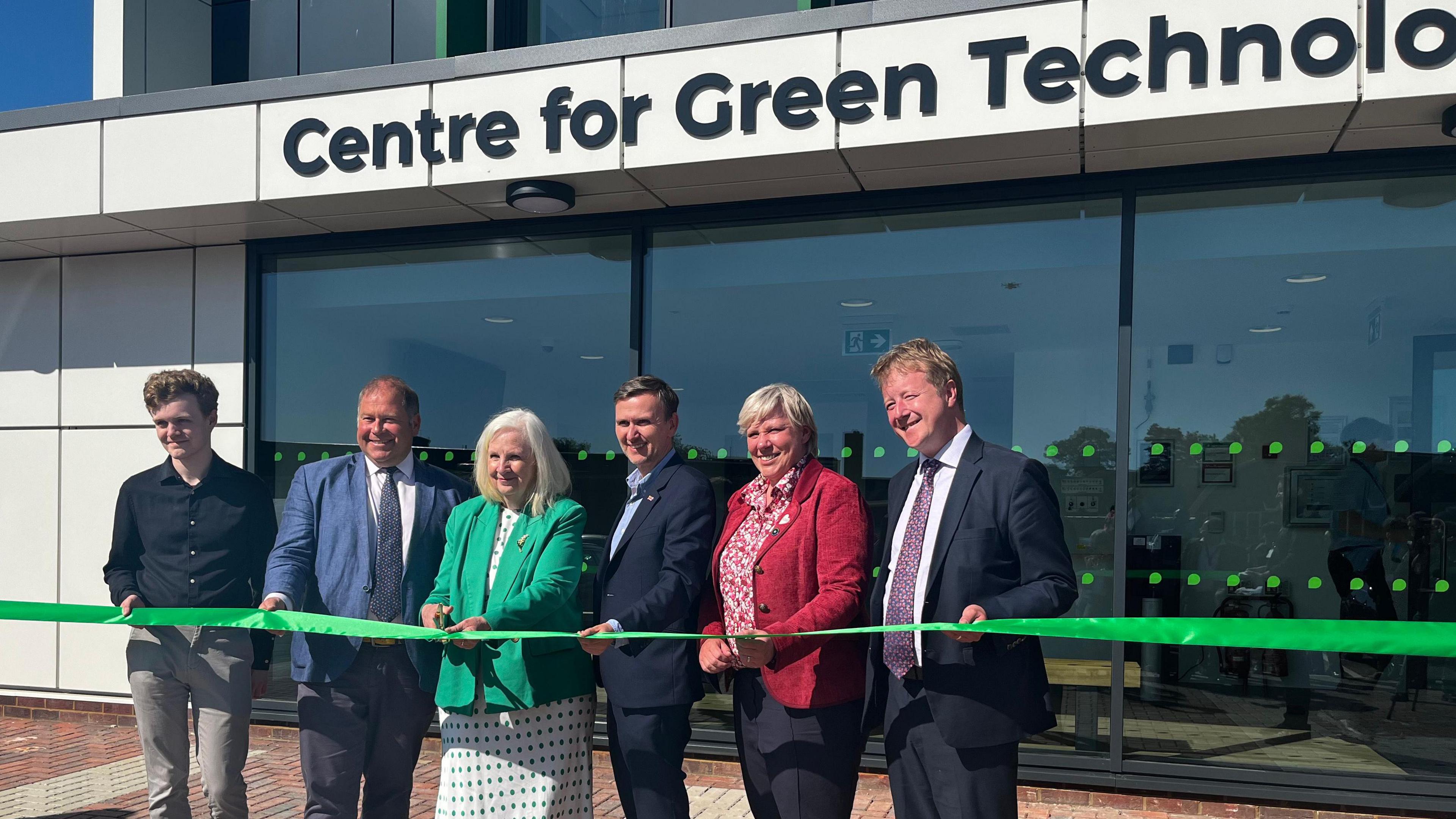A line of dignitaries at the opening of the centre. They are standing behind a green ribbon which a woman in a green jacket is about to cut with a pair of scissors.