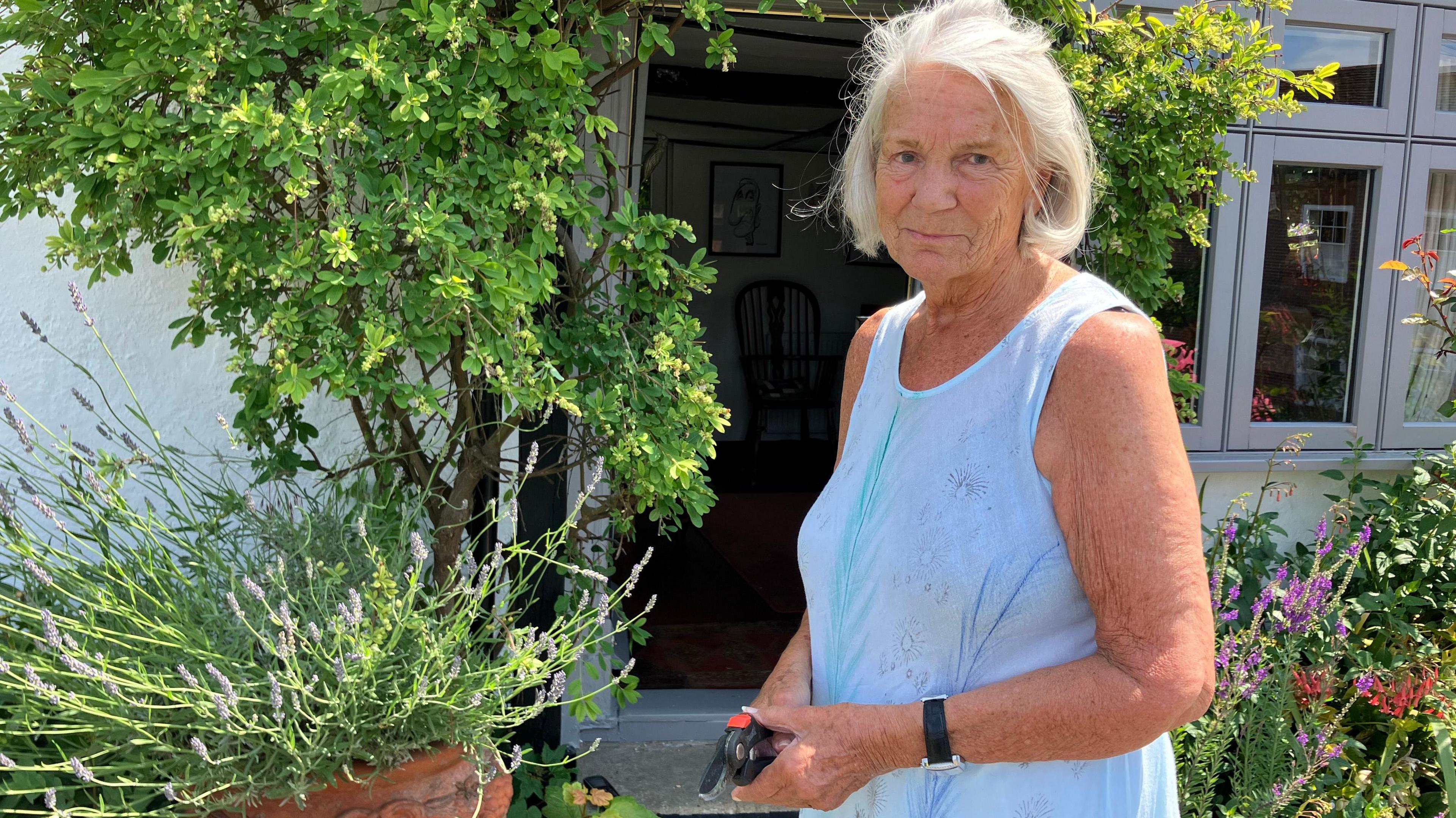 Christine Ashford-Hodges in a light blue dress, pruning her plants and potted trees, but looking at the camera