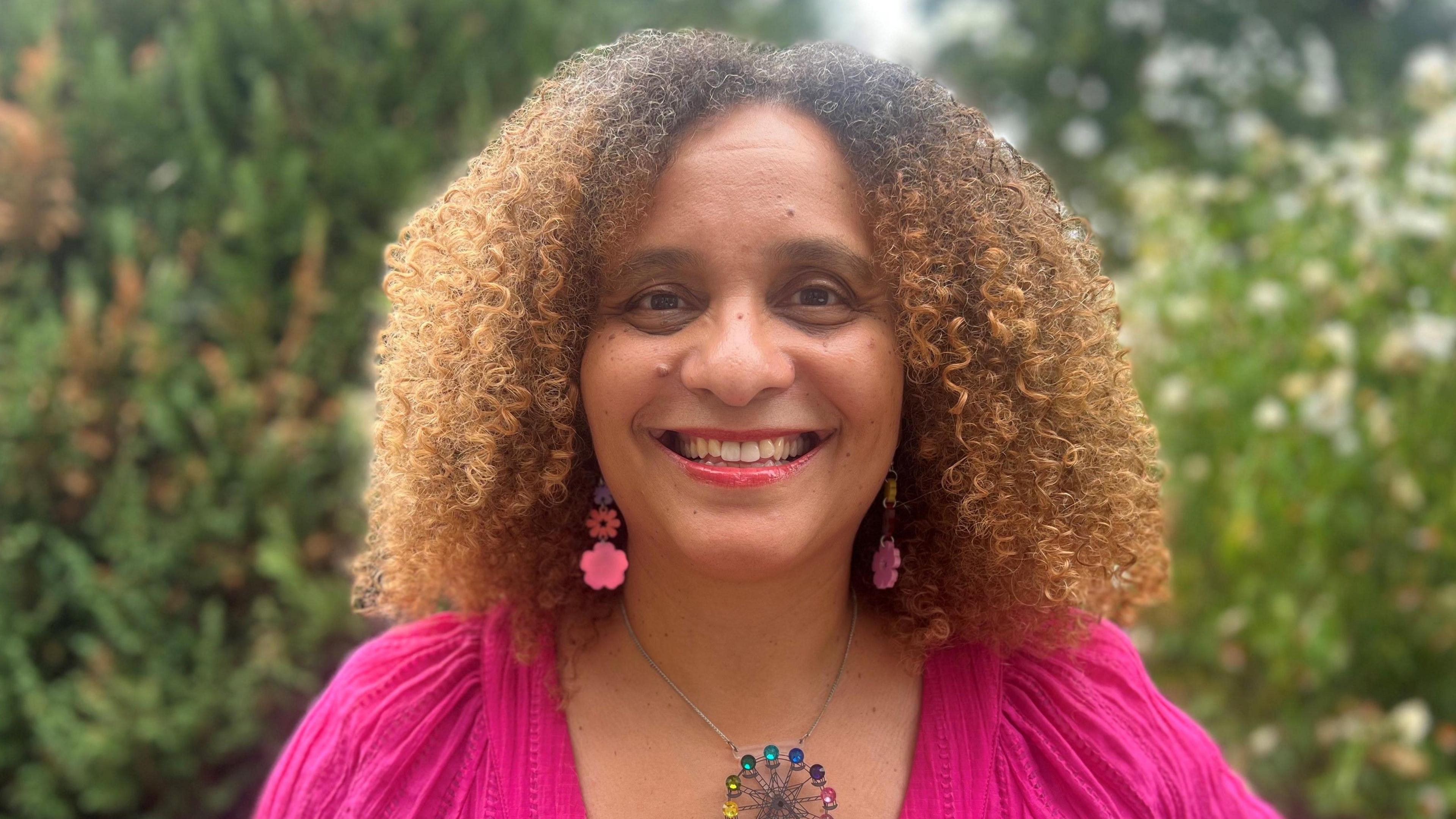 Susan Cole, who has tight curly brown hair, is wearing a pink top and pink dangly earrings in the shape of flowers and a colourful necklace. She is smiling at the camera.