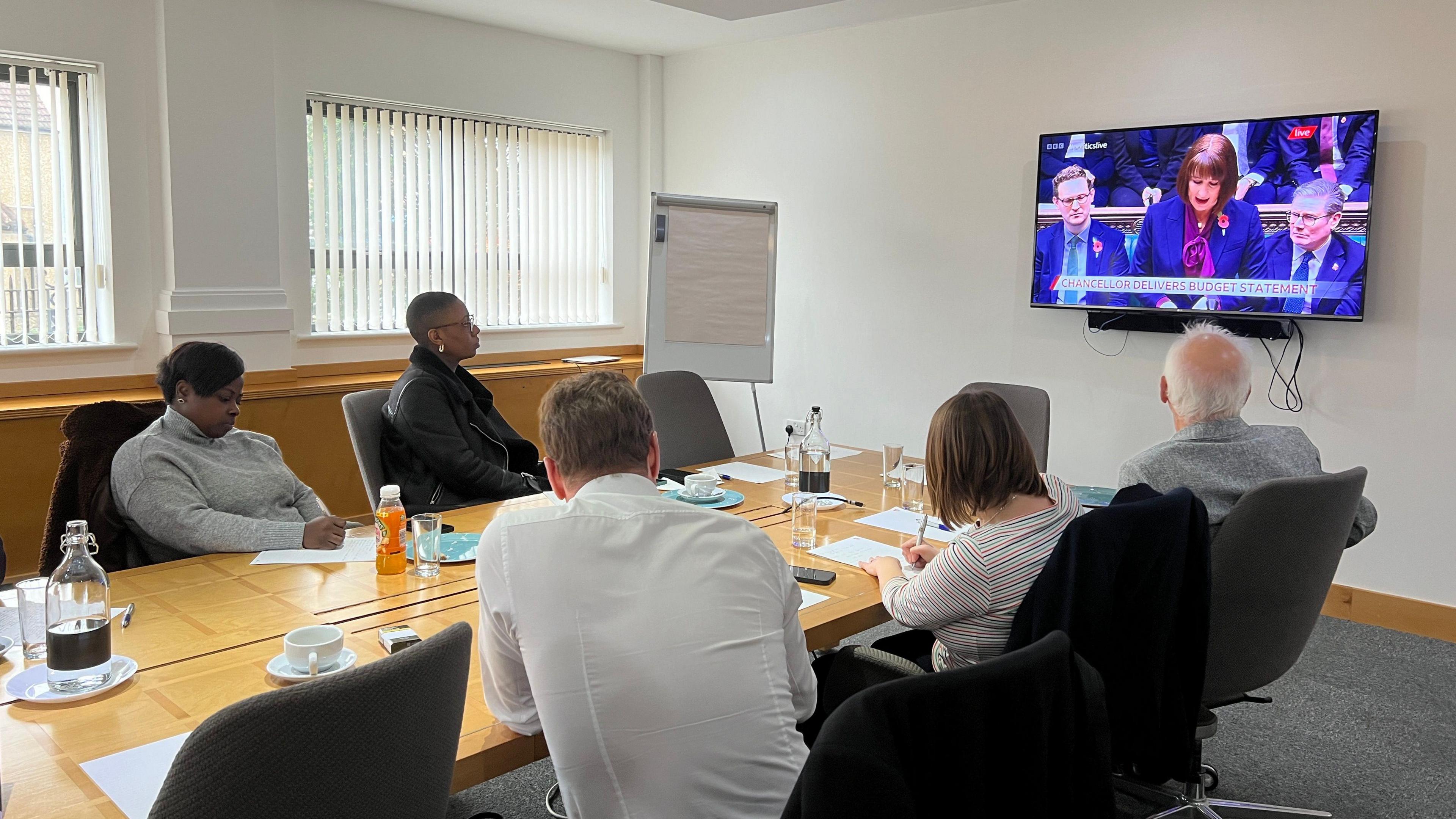 Five people sat around a conference table with refreshments in front of them. They are all watching a TV that shows Rachel Reeves as she stands at the Despatch Box in Parliament and delivers the budget. There is a whiteboard to the right of the screen. 