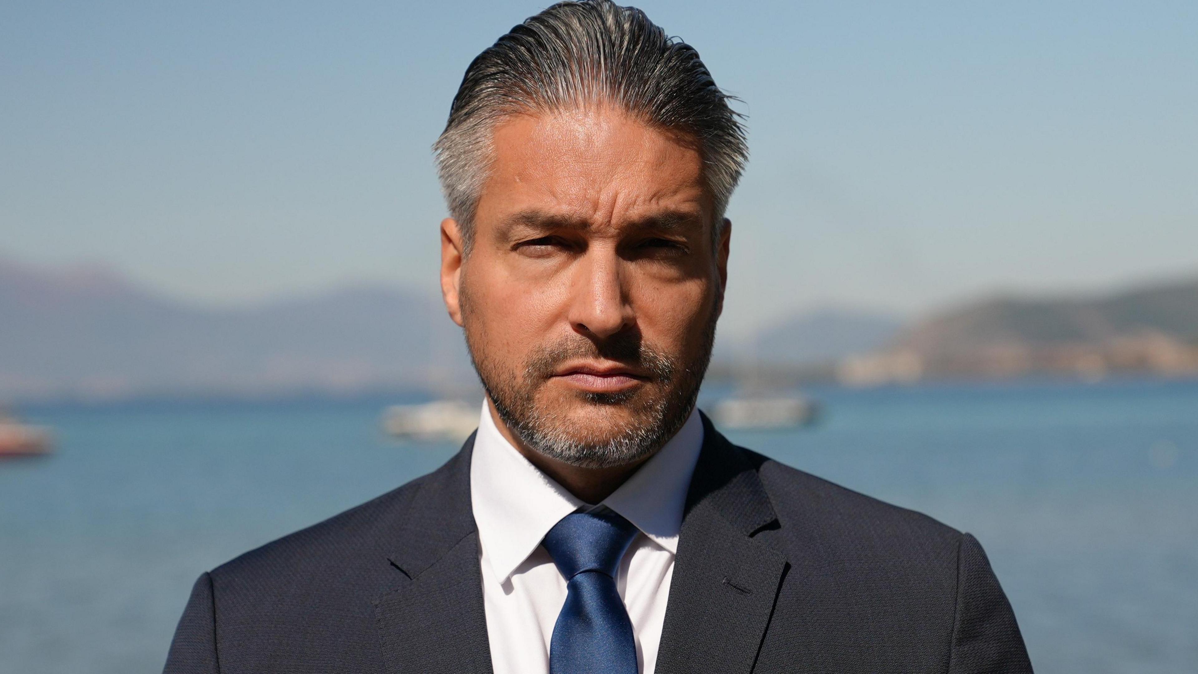 Mark Donfried, director of the Academy for Cultural Diplomacy, wearing a navy suit, white shirt and navy tie, on the shores of Lake Ohrid