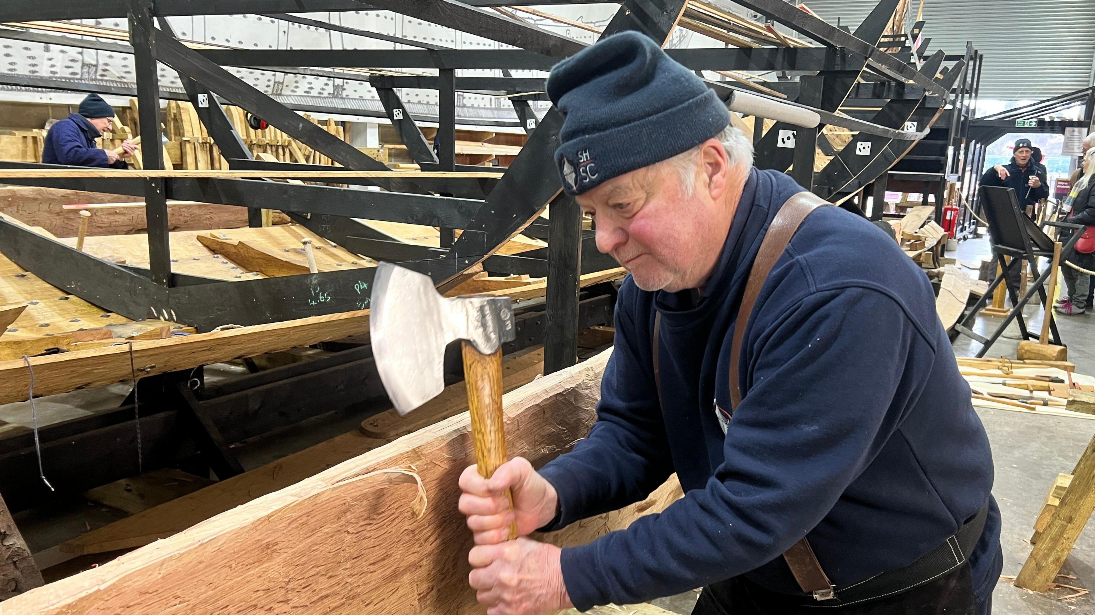 A volunteer holds an axe next to a plank of wood he aims to chop for the project. He  wears a navy jumper as well as a navy beanie hat.