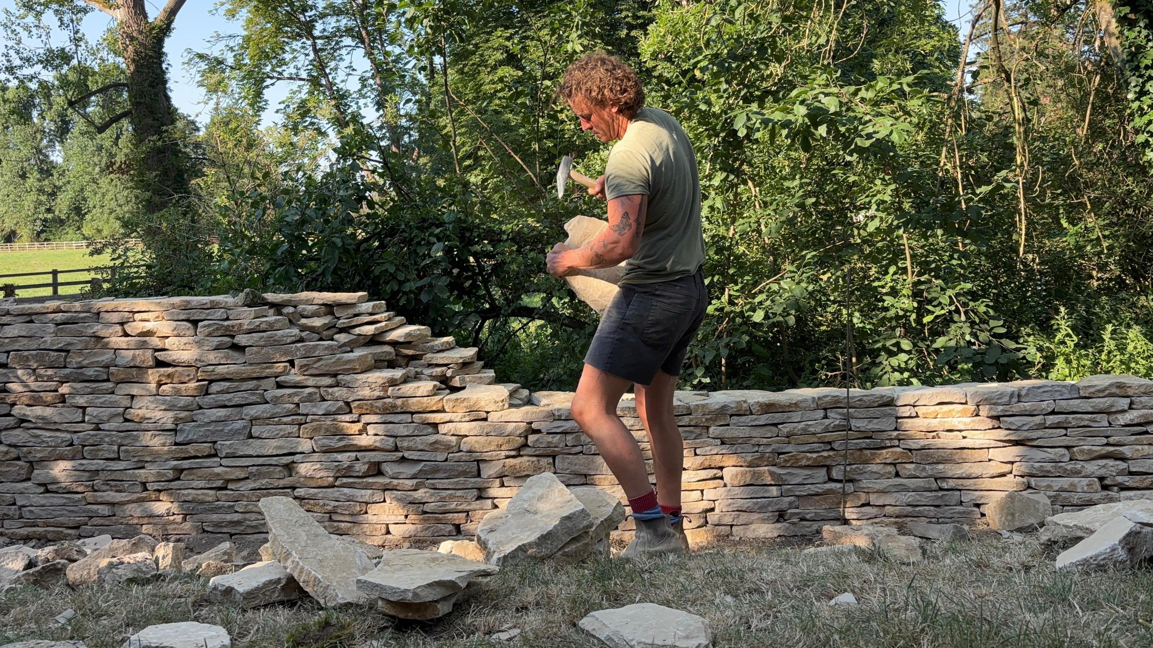 Joe Hanlon wearing black shorts and a green t-shirt. He is using a small sledgehammer to break up a large section of stone to use on a dry stone wall, which is half finished behind him. 