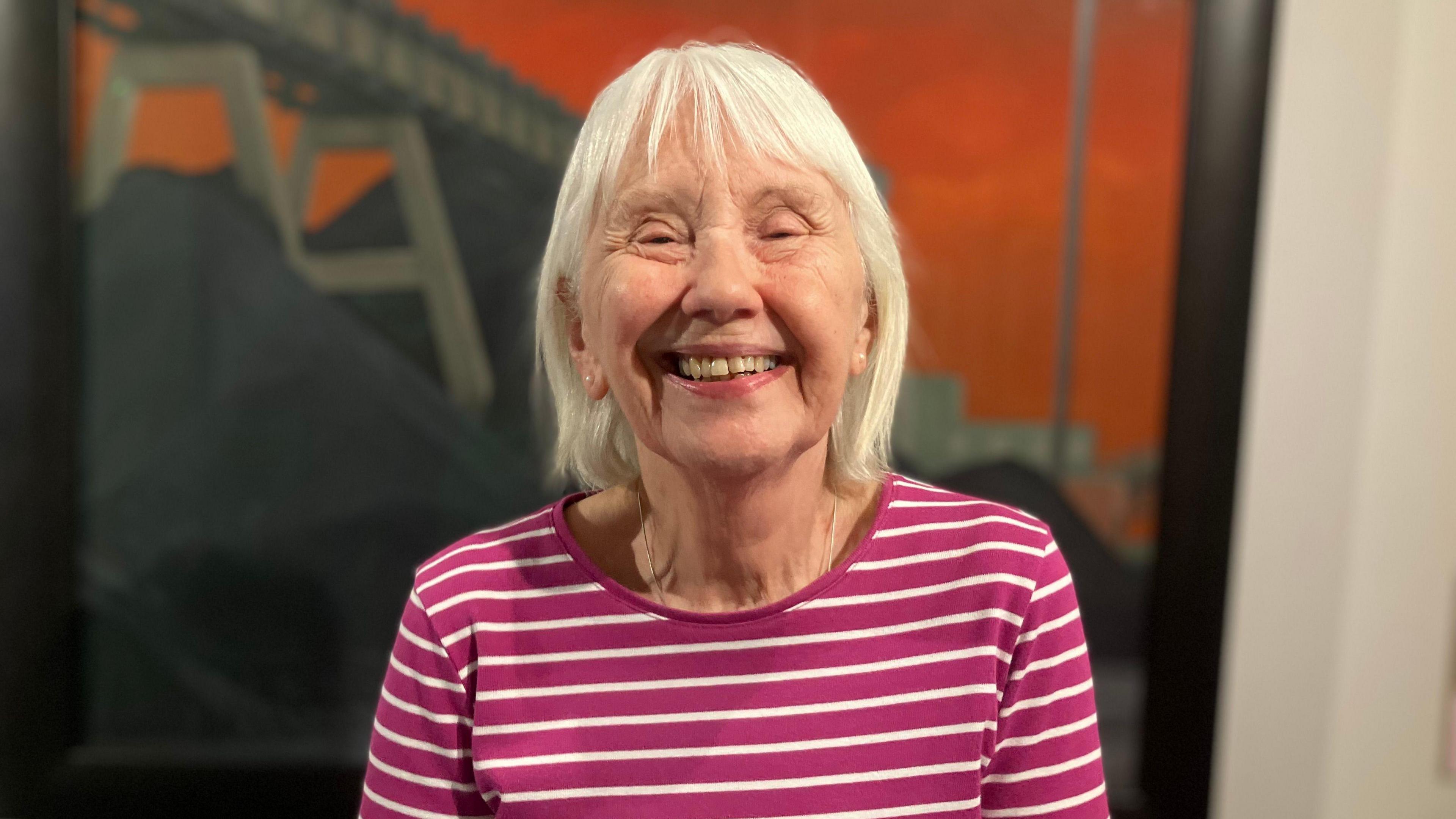 A woman with white hair smiles at the camera. She wears a red and white top. Behind her, a orange painting hung from the walls is blurred.