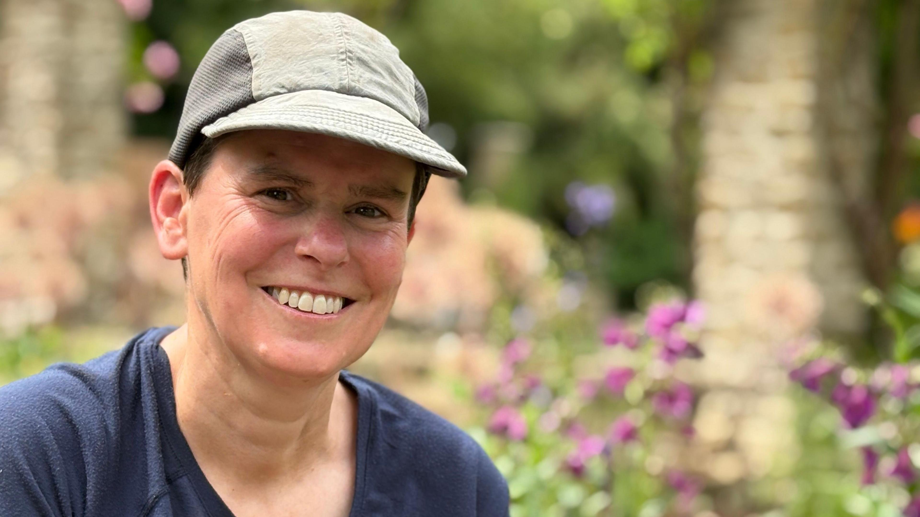 A women in her fifties wearing a faded white baseball cap smiling next to purple flowers