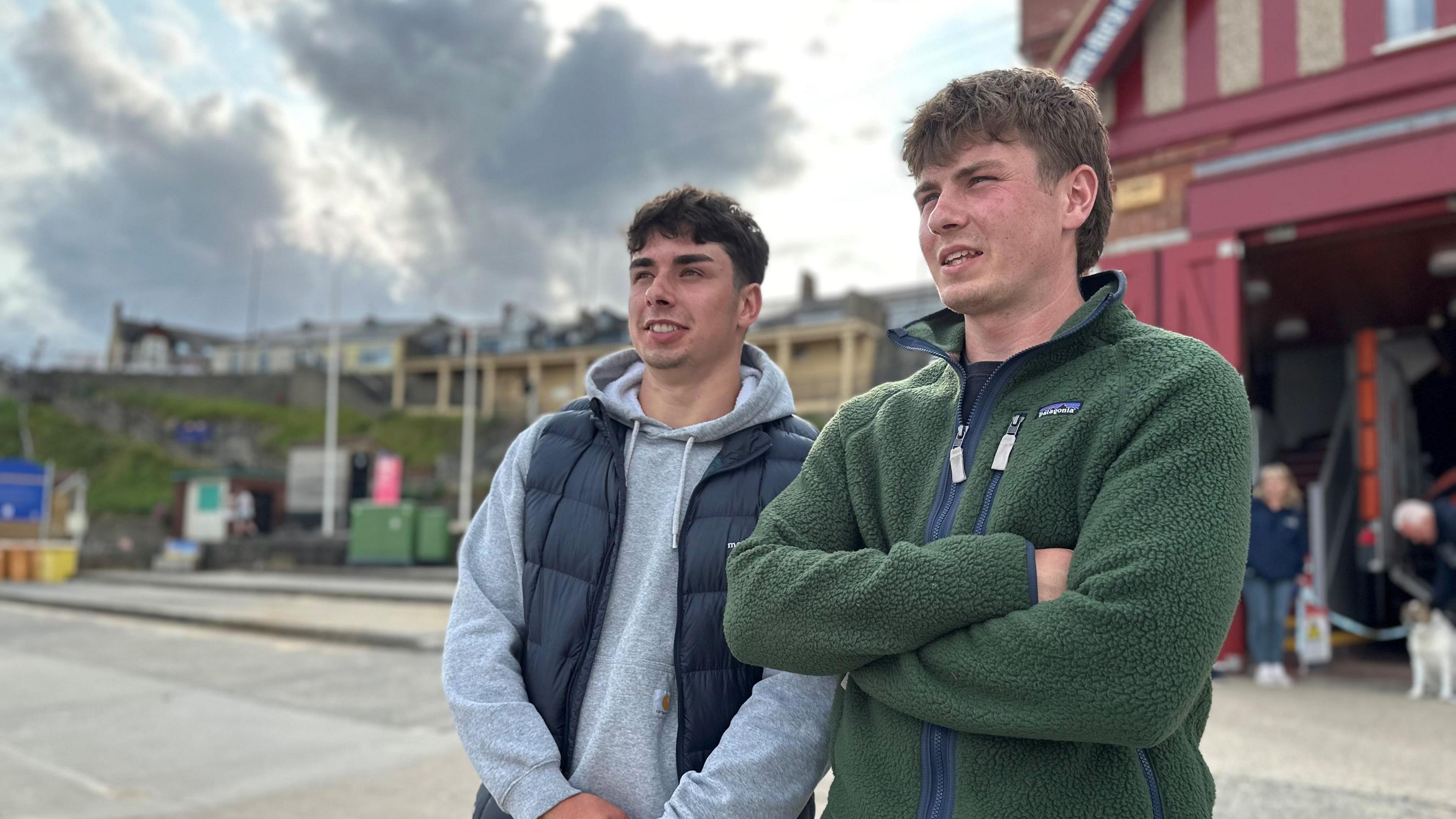 The two brothers stand side by side in front of the Cullercoats RNLI station, a large red and grey building, they are looking out to sea.