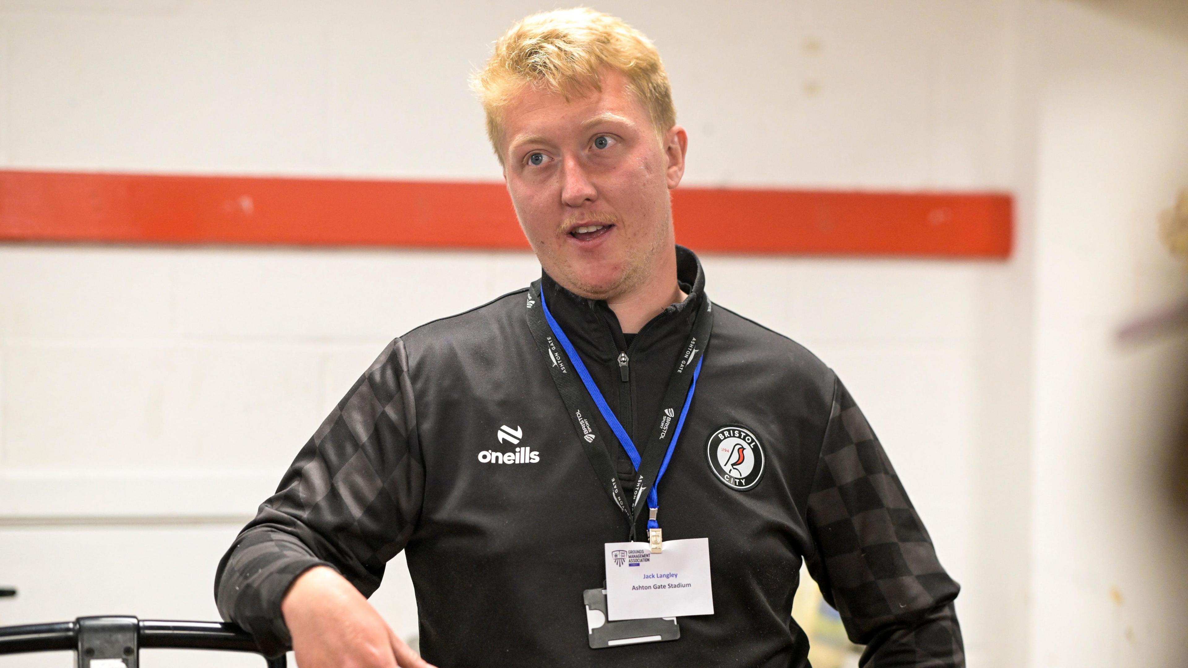 Jack Langley, wearing a black top and a lanyard with his name on it. He is a middle-aged man with ginger hair, and is pictured in a dressing room mid-conversation