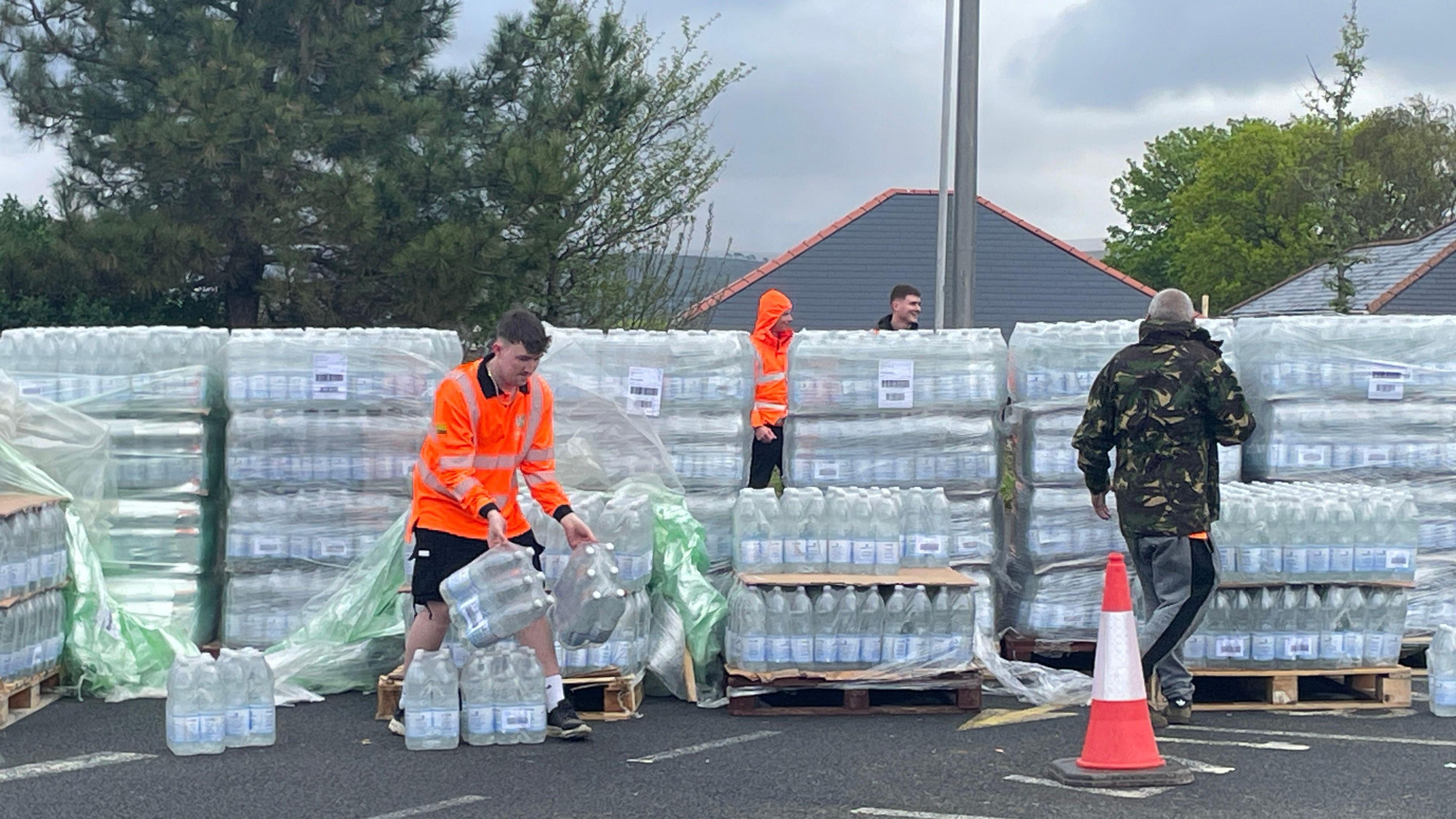 A man wearing an orange hi-vis jacket moving packs of bottled water. Behind him is packs of bottled water stacked on wooden crates, with a person wearing a camouflage coat and grey trousers to the right, and people stood behind stack of water bottles.