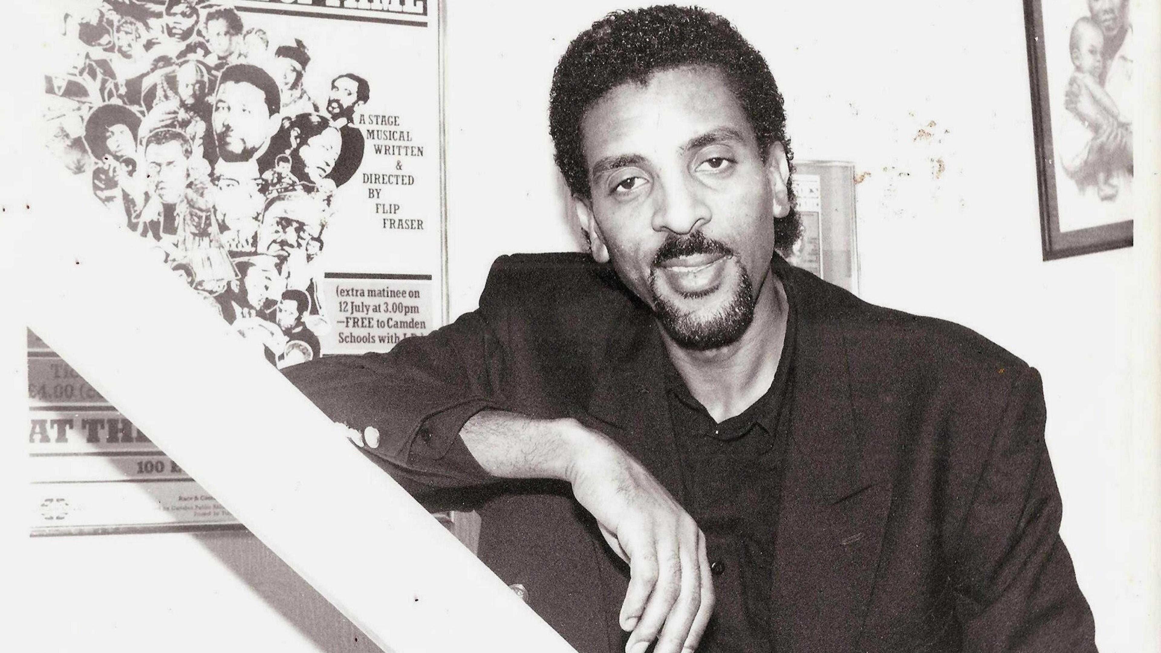 A black and white photo of Peter Randolph Fraser, a man who originally comes from Jamaica, who has dark hair and a goatee beard, wearing a dark shirt and suit. He stands in front of a poster.