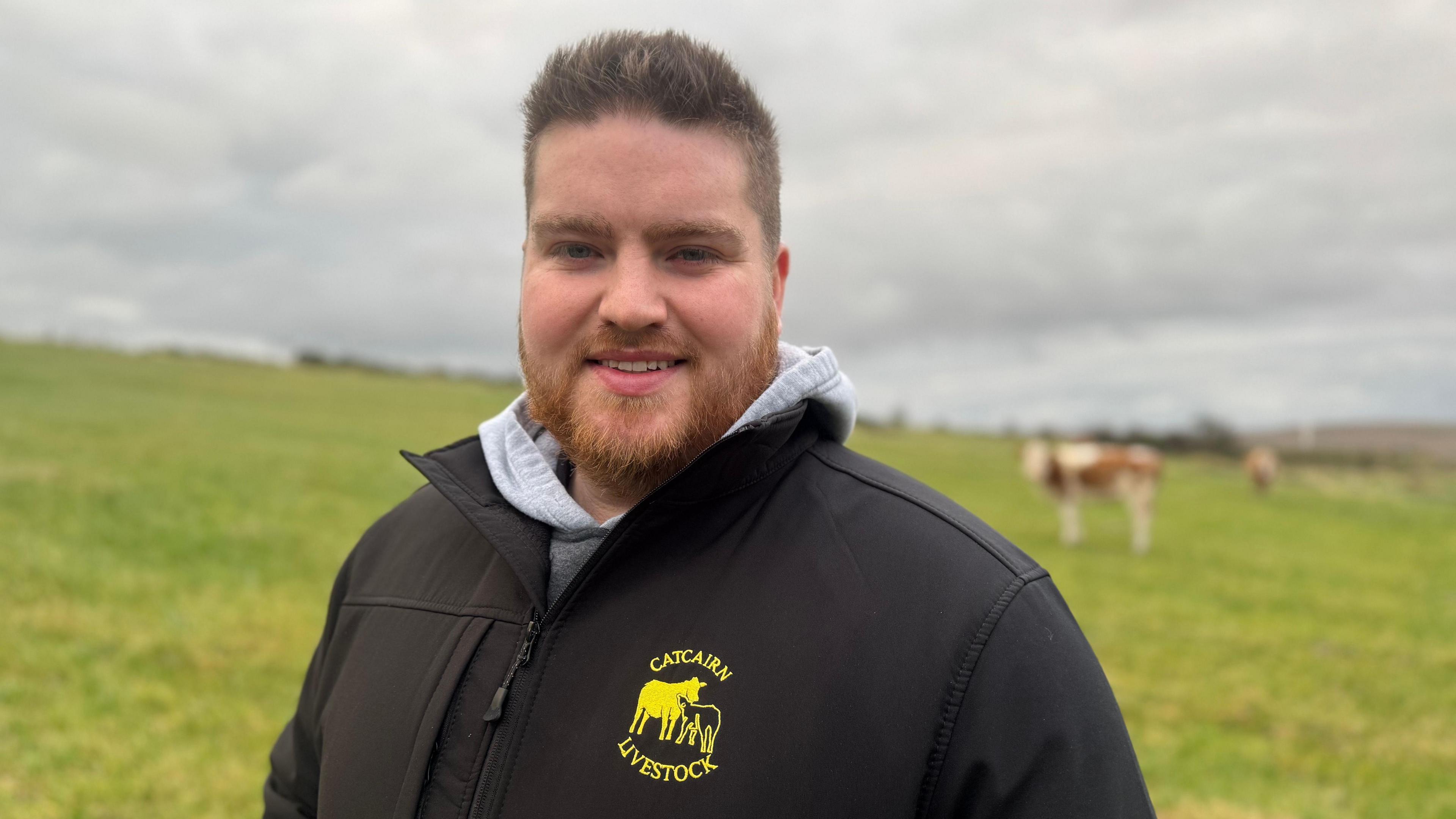 A man, standing in front of a field of grass, looking at the camera. He is pictured from the chest up. He is wearing a grey hoodie and a black jacket with a yellow logo with a cow on it. He has short brown hair and a tight beard. There is a cow in the background to the right of the man. The sky is full of grey clouds. 