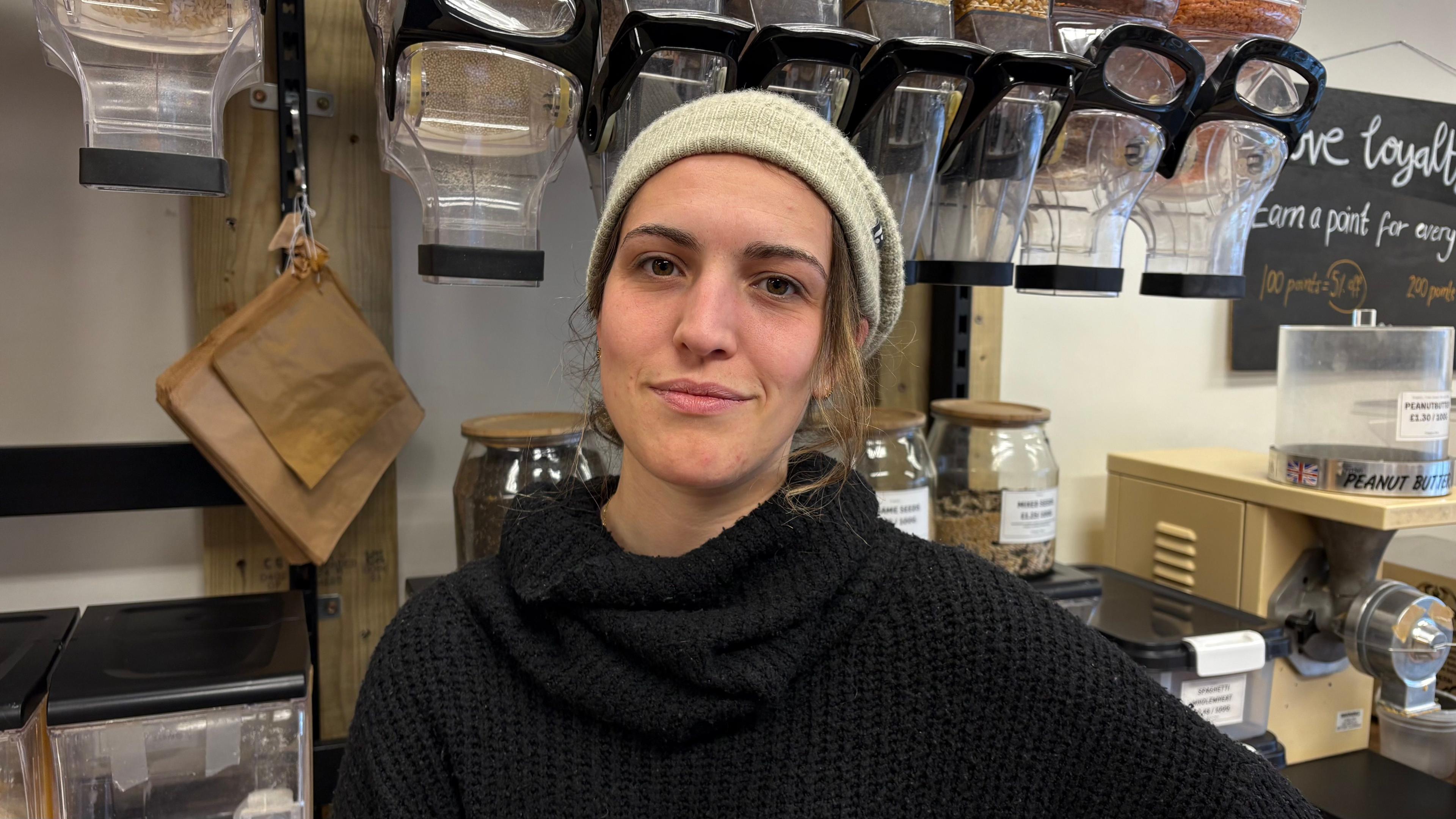 Heni Cloake with long light brown hair wearing a green beanie hat and a blue pullover with jars of refill food behind her. There is also a mincing machine behind and a blackboard advertising a loyalty scheme.