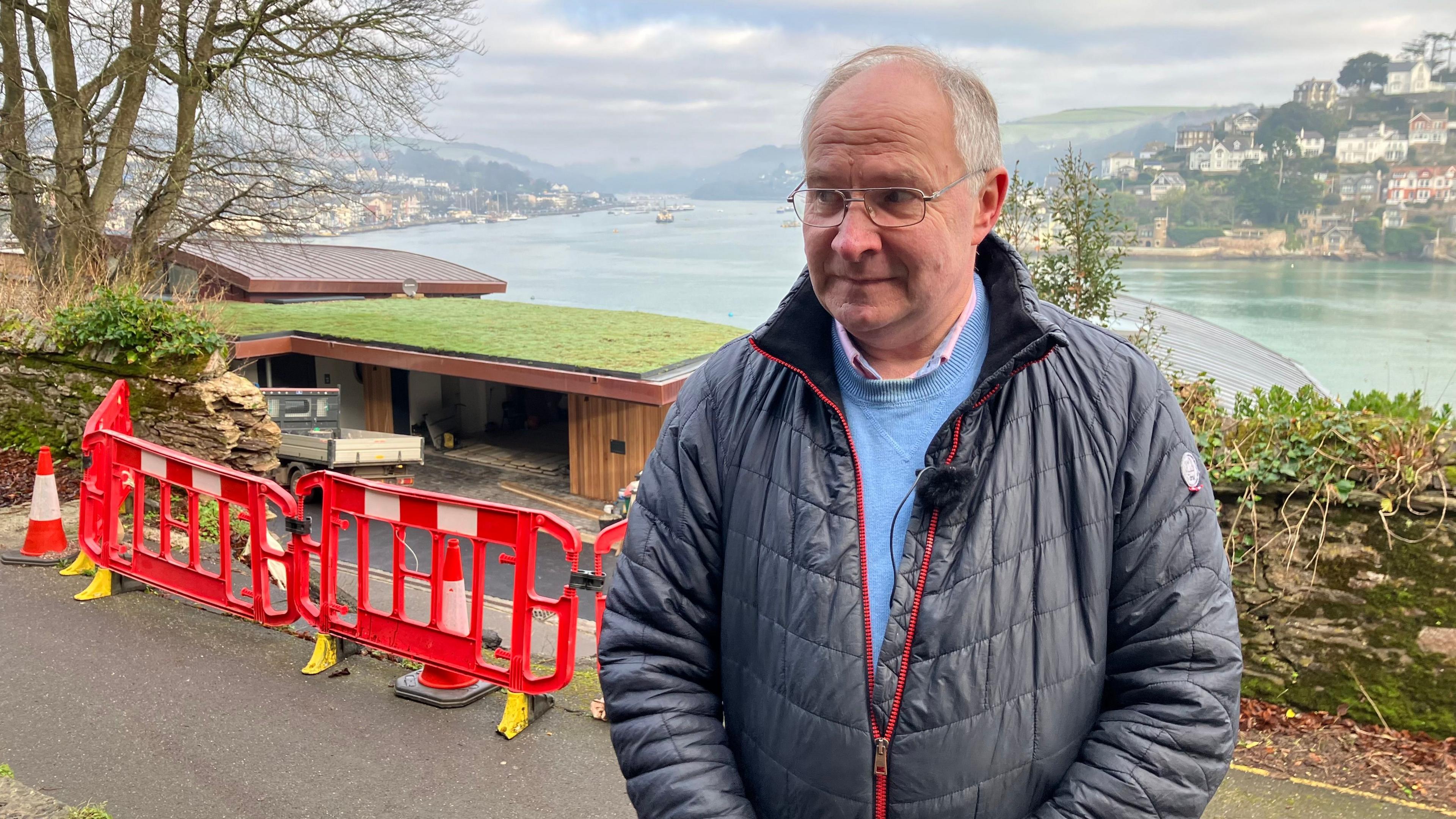 Cllr Jonathan Hawkins wearing glasses and looking to the left. He is wearing a light blue jumper with a black coat over the top. He is stood in front of the red barriers surrounding the landslide.