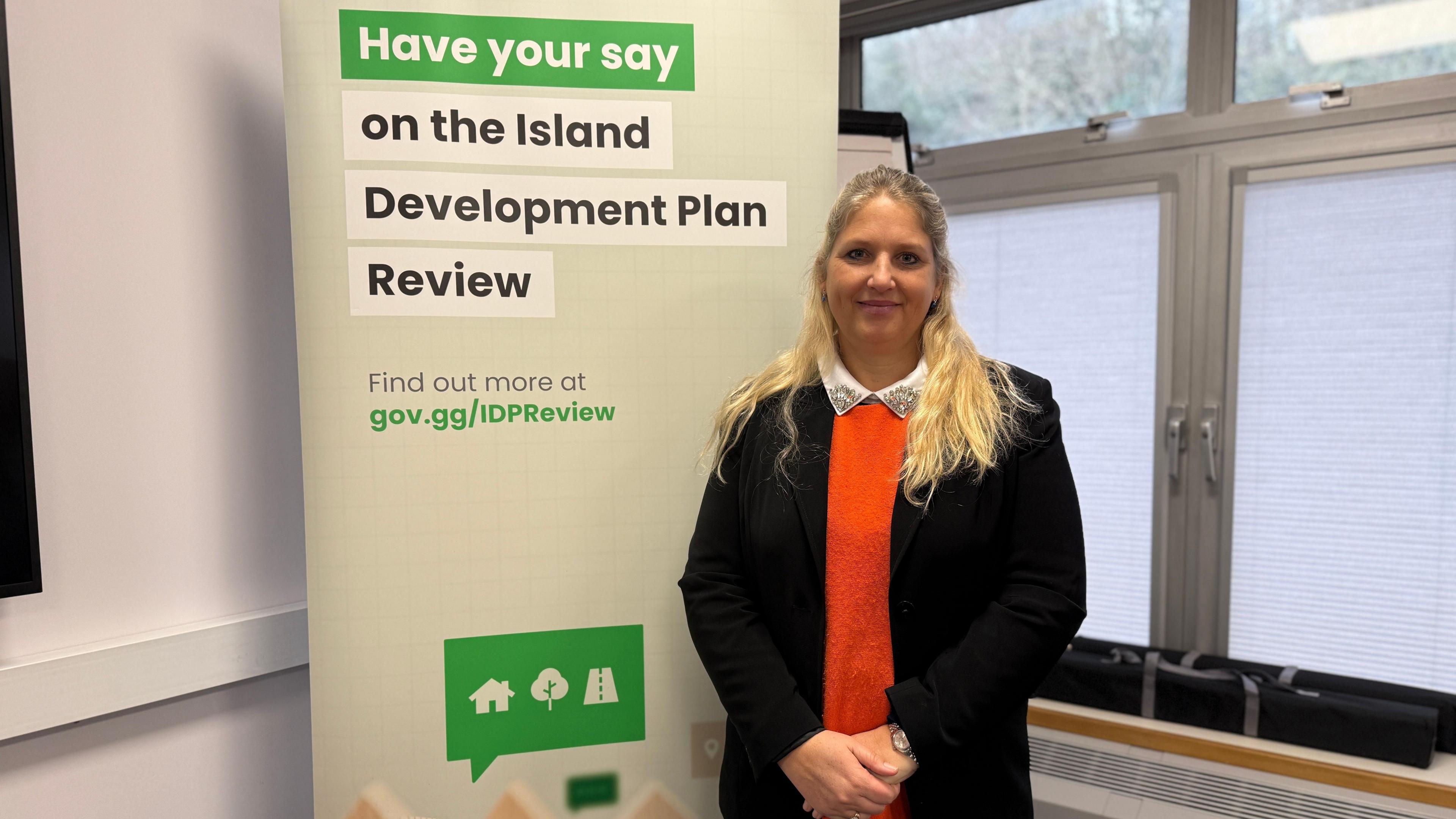 A picture of Deputy Victoria Oliver who is a women with long blonde hair. She is wearing an orange jumper with a black blazer. She is standing next to a sign which says 'Have your say on the Island Development Plan Review'.