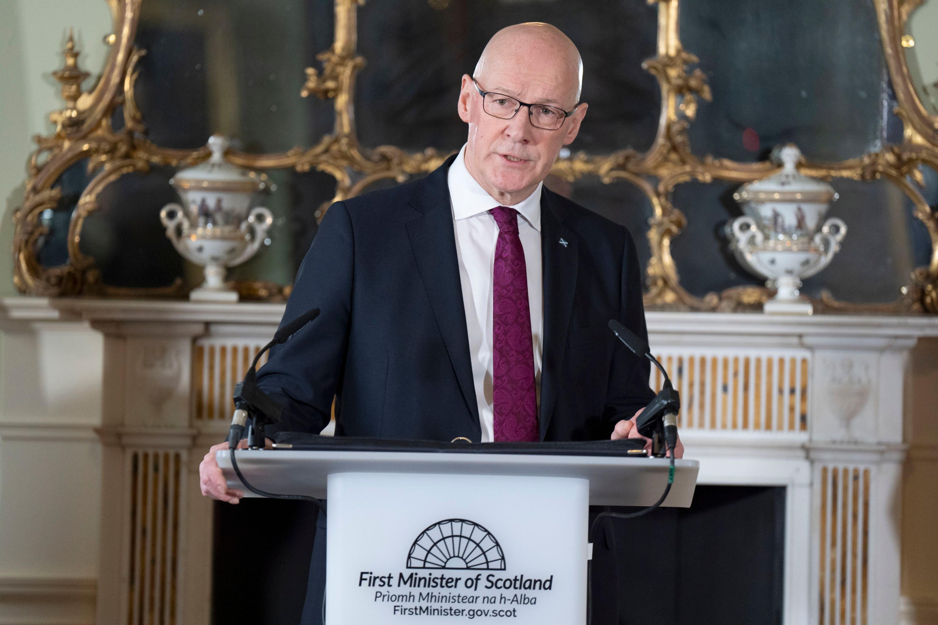 John Swinney stands at the pulpit in Bute House, standing in front of a fireplace with an ornate mirror and vases. He is wearing a navy blue suit, a white shirt and a purple tie.