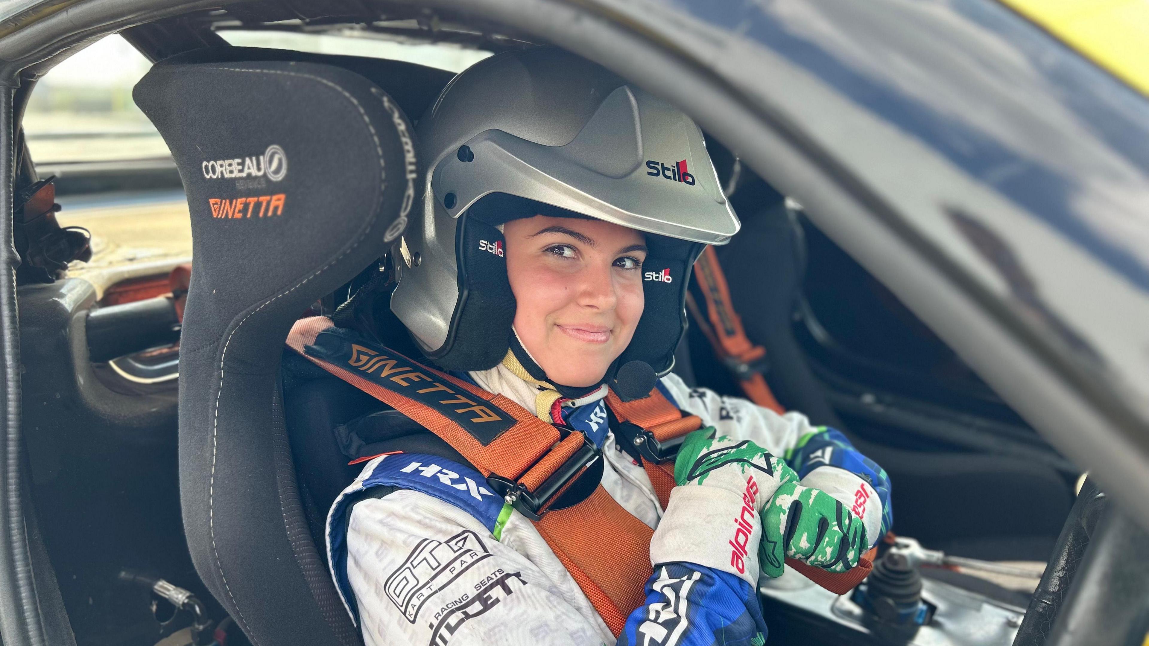 A young girl, 14 sat in a G40, wearing an orange harness - and a grey helmet. She's wearing gloves and a race suit behind the steering wheel about to set off around the track.