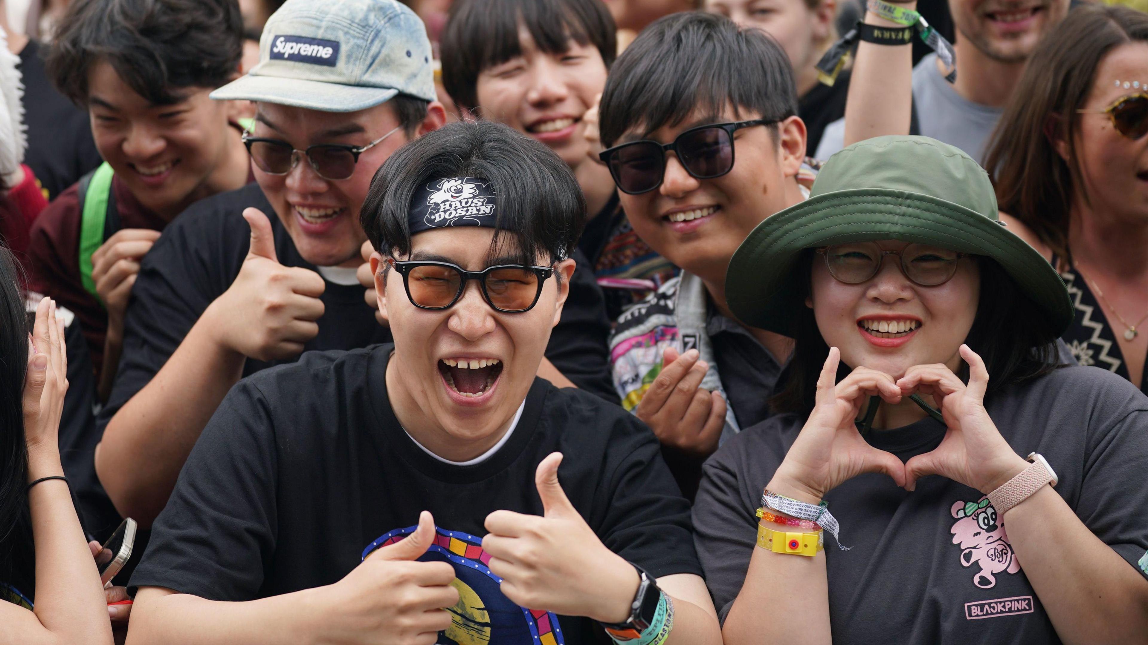 Image of audience members at Glastonbury Festival. Three men can be smiling with their thumbs up, and a woman is making a heart sign with her hands.