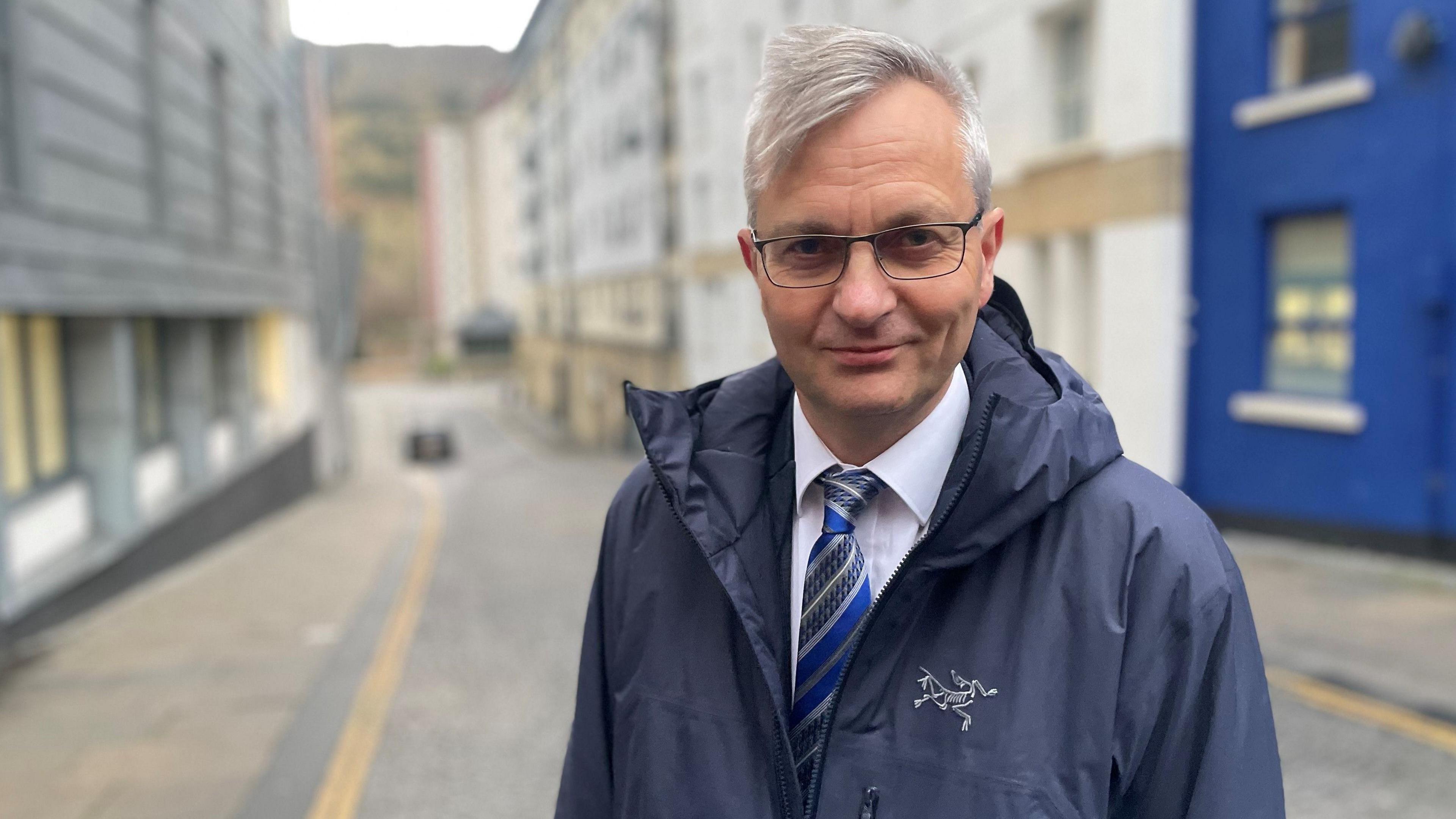 Stephen Sandham wearing a blue jacket over a white shirt and a blue and grey striped tie. He has grey hair swept to the right side and glasses with a dark-coloured frame. He is standing in front of a blurred background.