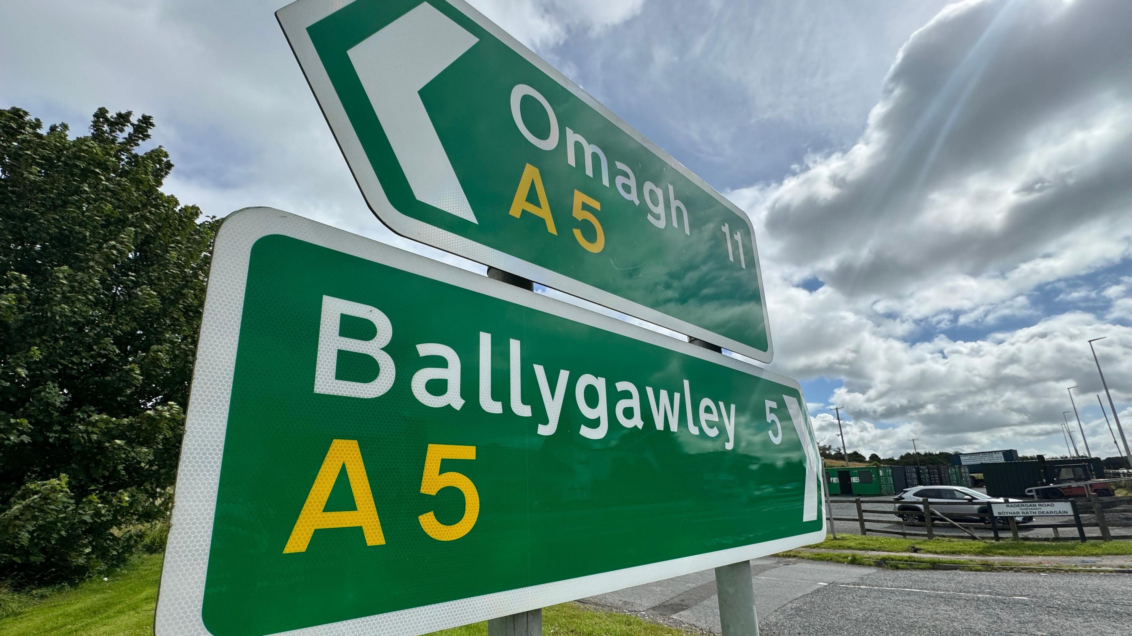 Two green road signs, one reading Omagh A5 and the other reading Ballygawley A5. There is a cloudy sky above.