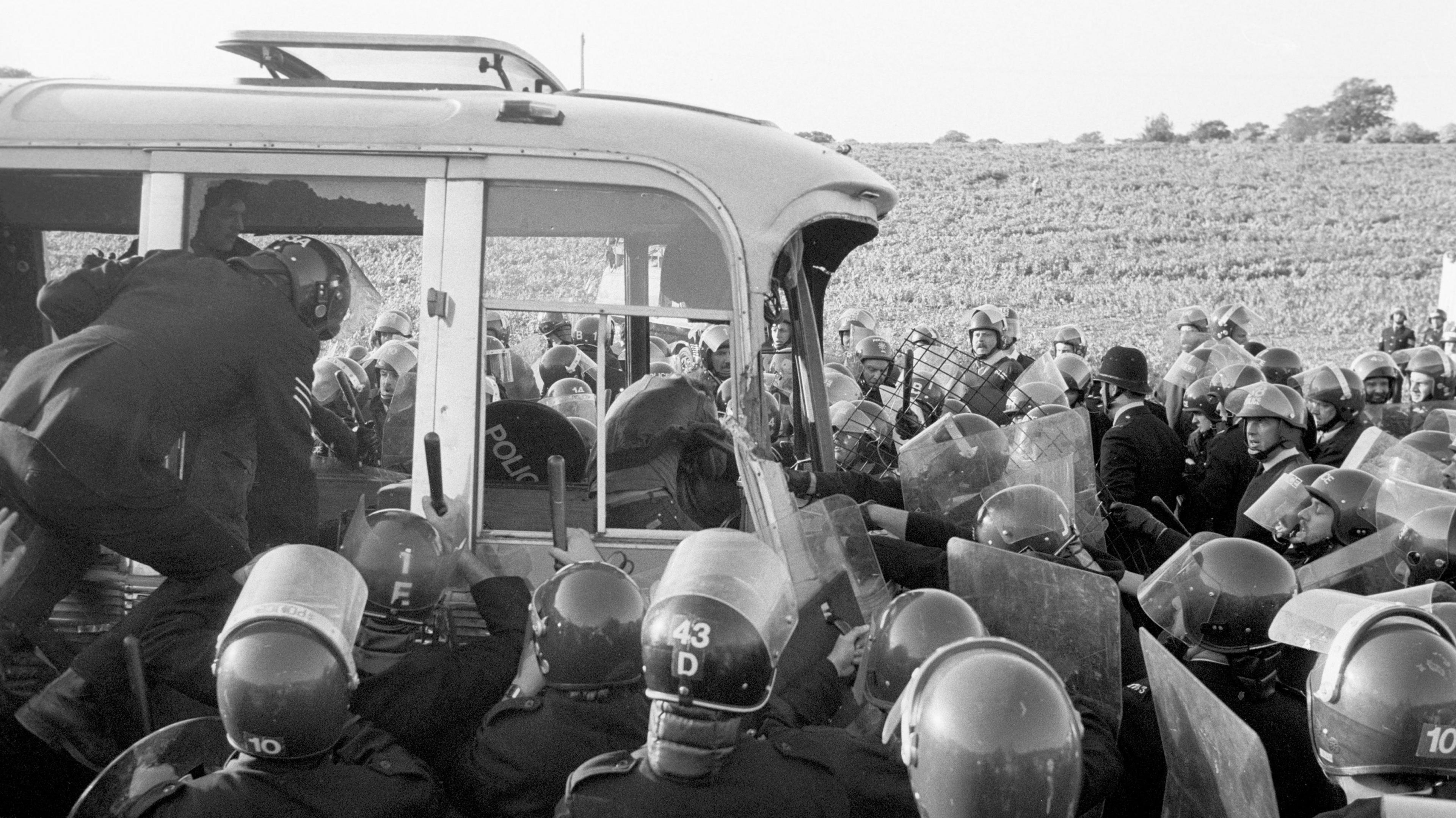 A black & white photo of police in riot gear surrounding an old bus