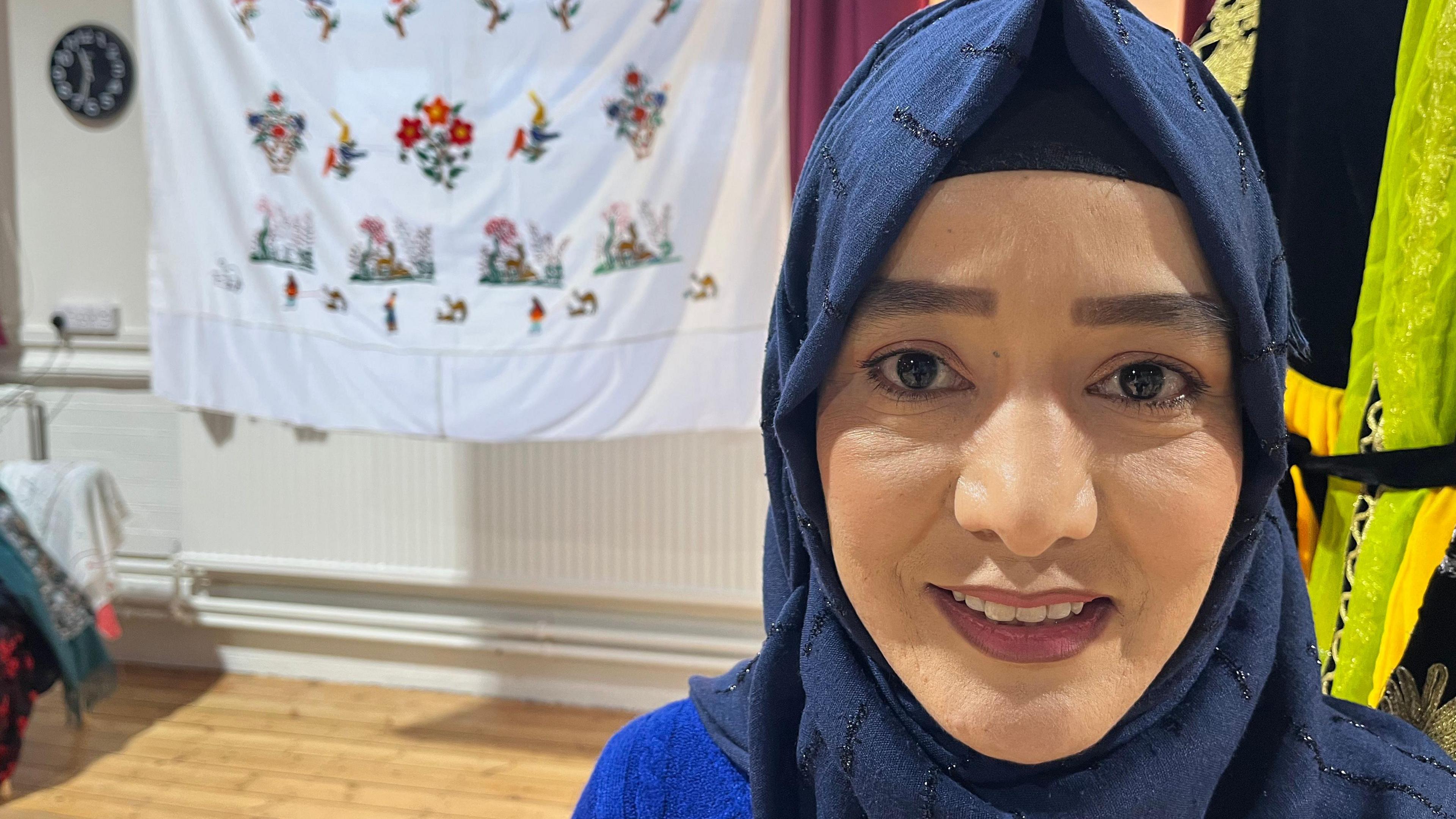 lady in hijab faces the camera with some needlework her mother created behind her 
