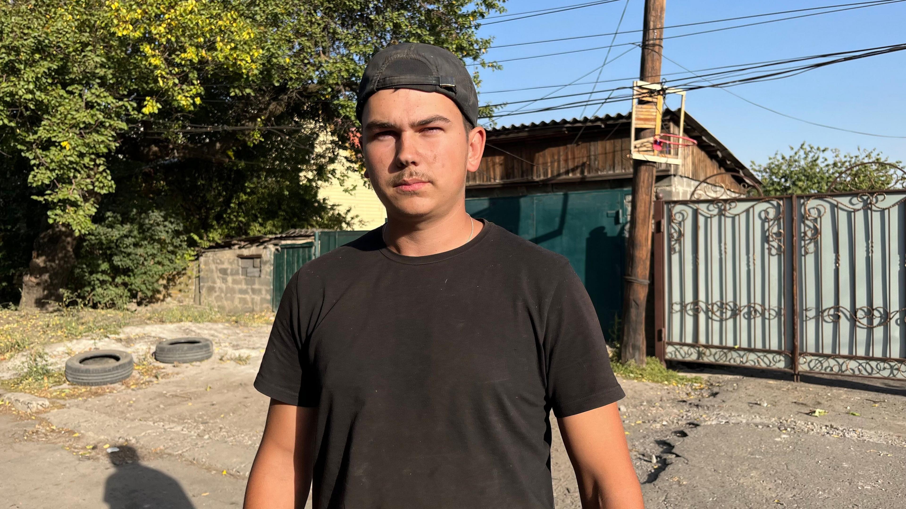 Roman stands in the street, in front of a garage and some gates. He has a feint moustache, and is wearing a black T-shirt and a black baseball cap the wrong way round.