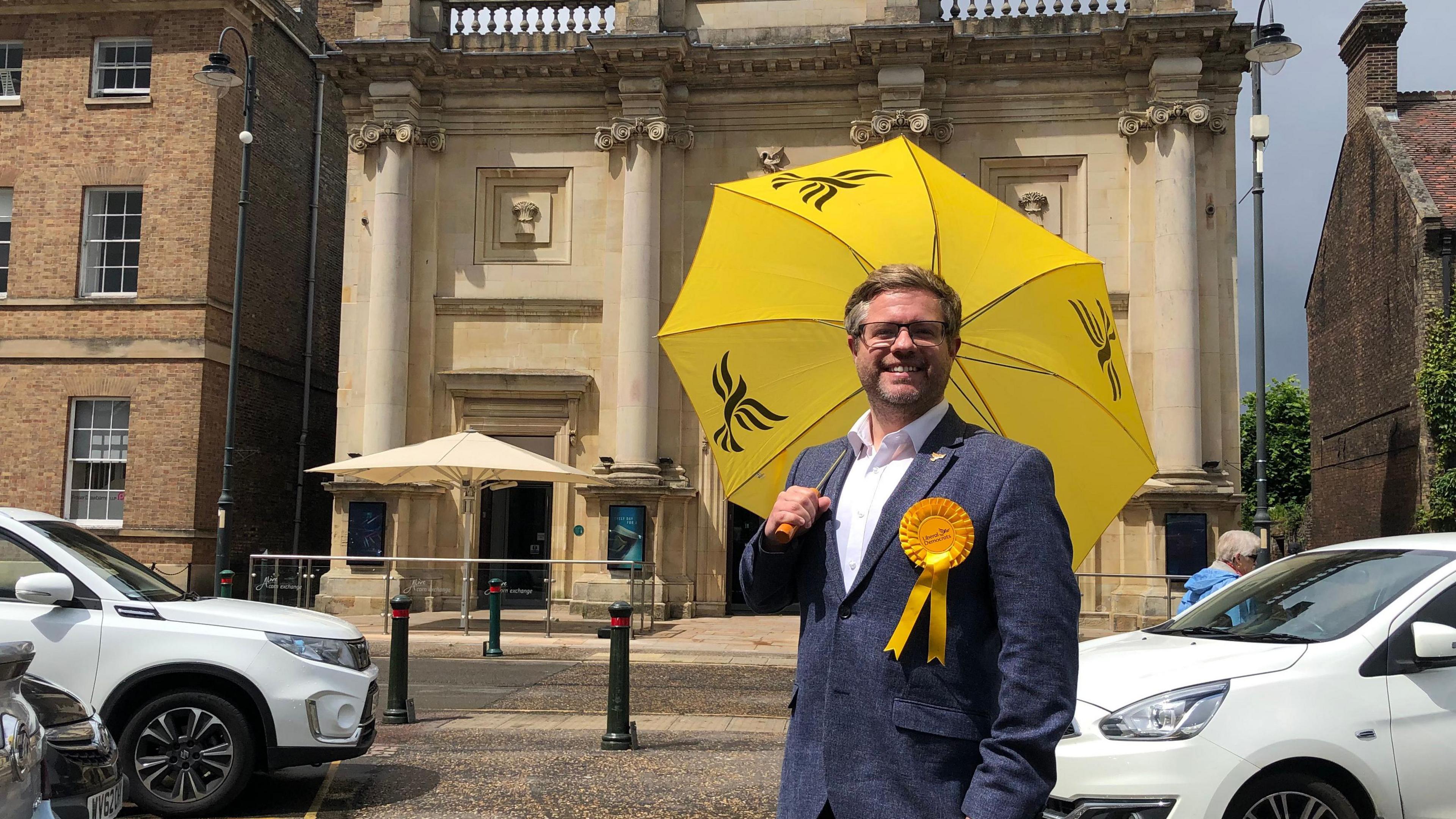 Liberal Democrat candidate for North West Norfolk stands on the Tuesday Market Place in King's Lynn holding a bright yellow Lib Dem branded umbrella with a yellow rosette pinned to his blue suit