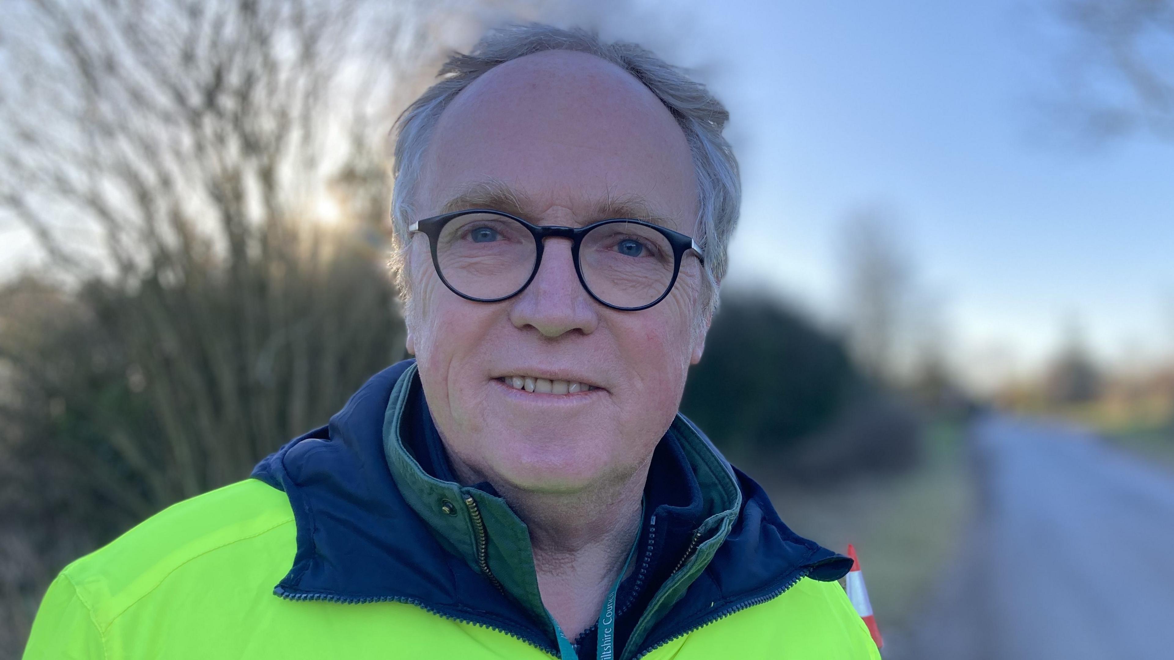 Brian Mathew MP for Melksham and Devizes wearing black-rimmed glasses and wearing a high-visibility jacket standing next to a road