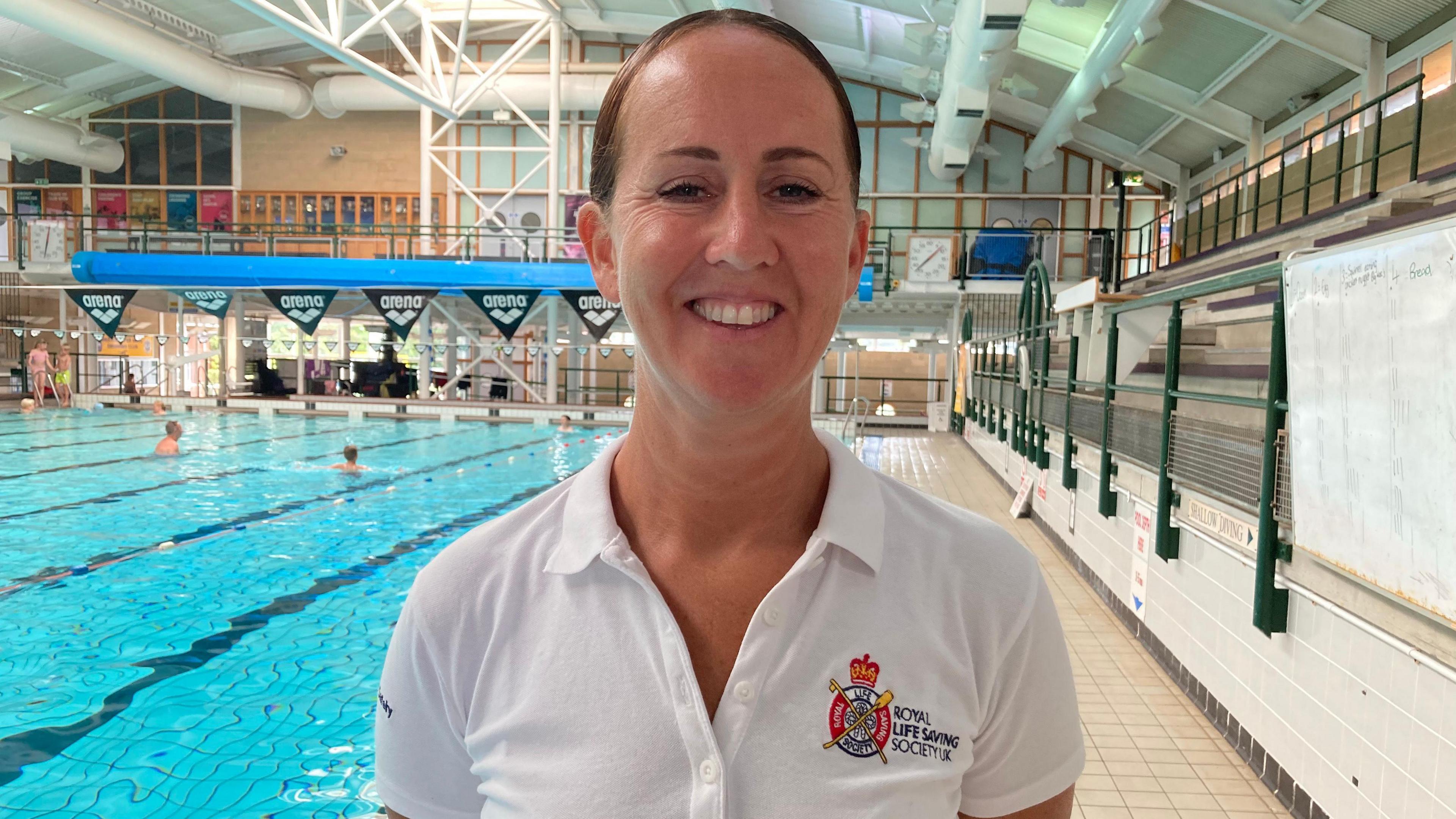 Jo Talbot. She has dark hair which is tied back. She is pictured in front of a swimming pool and is smiling, looking directly at the camera. She is wearing a white shirt with a royal lifesaving society UK logo on it. 