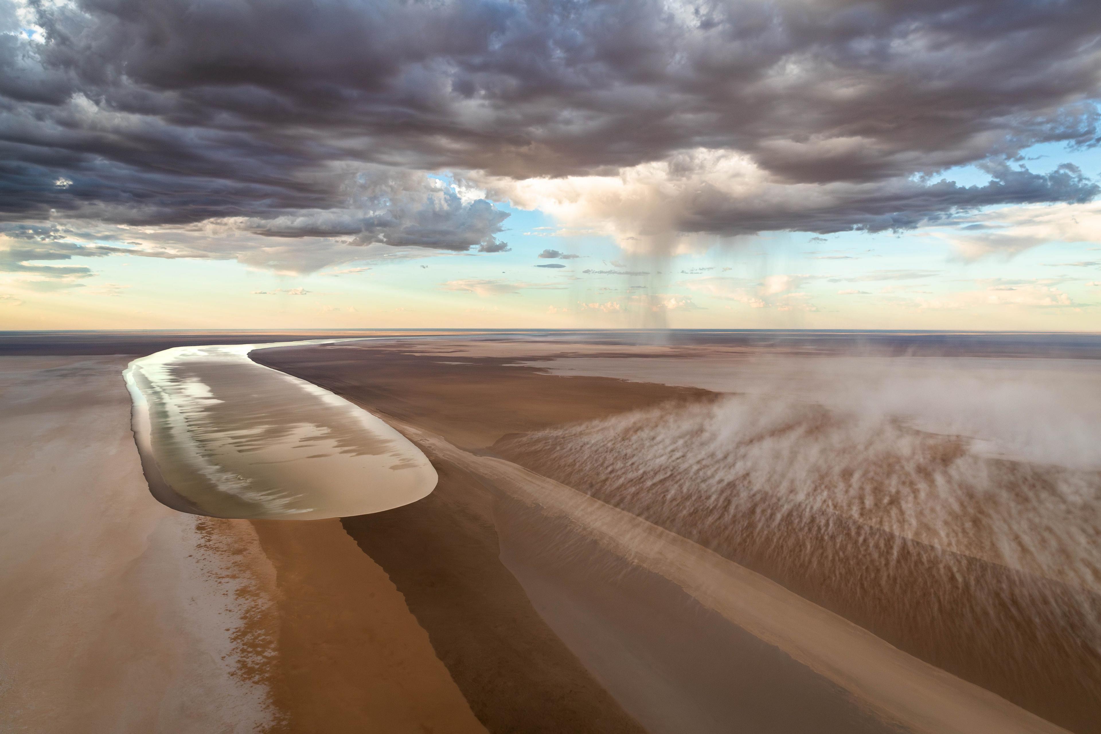 Aerial view of floodwaters carving through the cracked earth of Australia’s outback. Ribbons of water snake toward the vast salt flats of Kati Thanda-Lake Eyre.