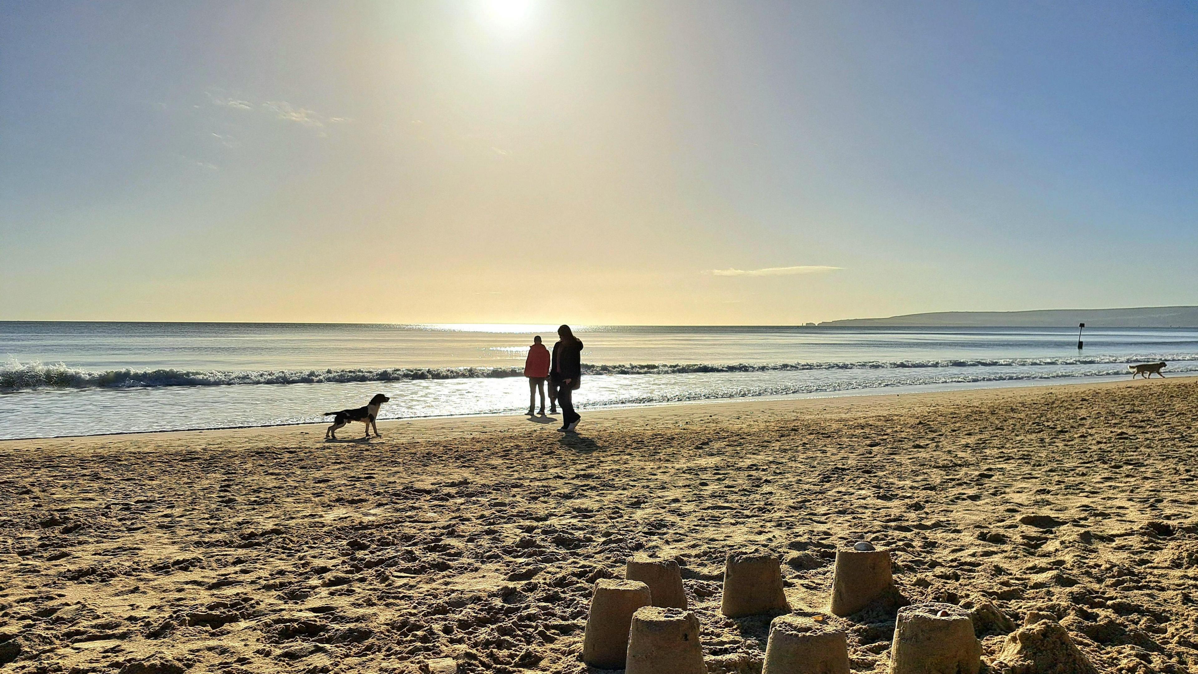 Two walkers are on a sandy beach as the sun is low in the sky. There is a small wave washing towards the sand. In the foreground you can see sandcastles. There are two dogs on the beach. 