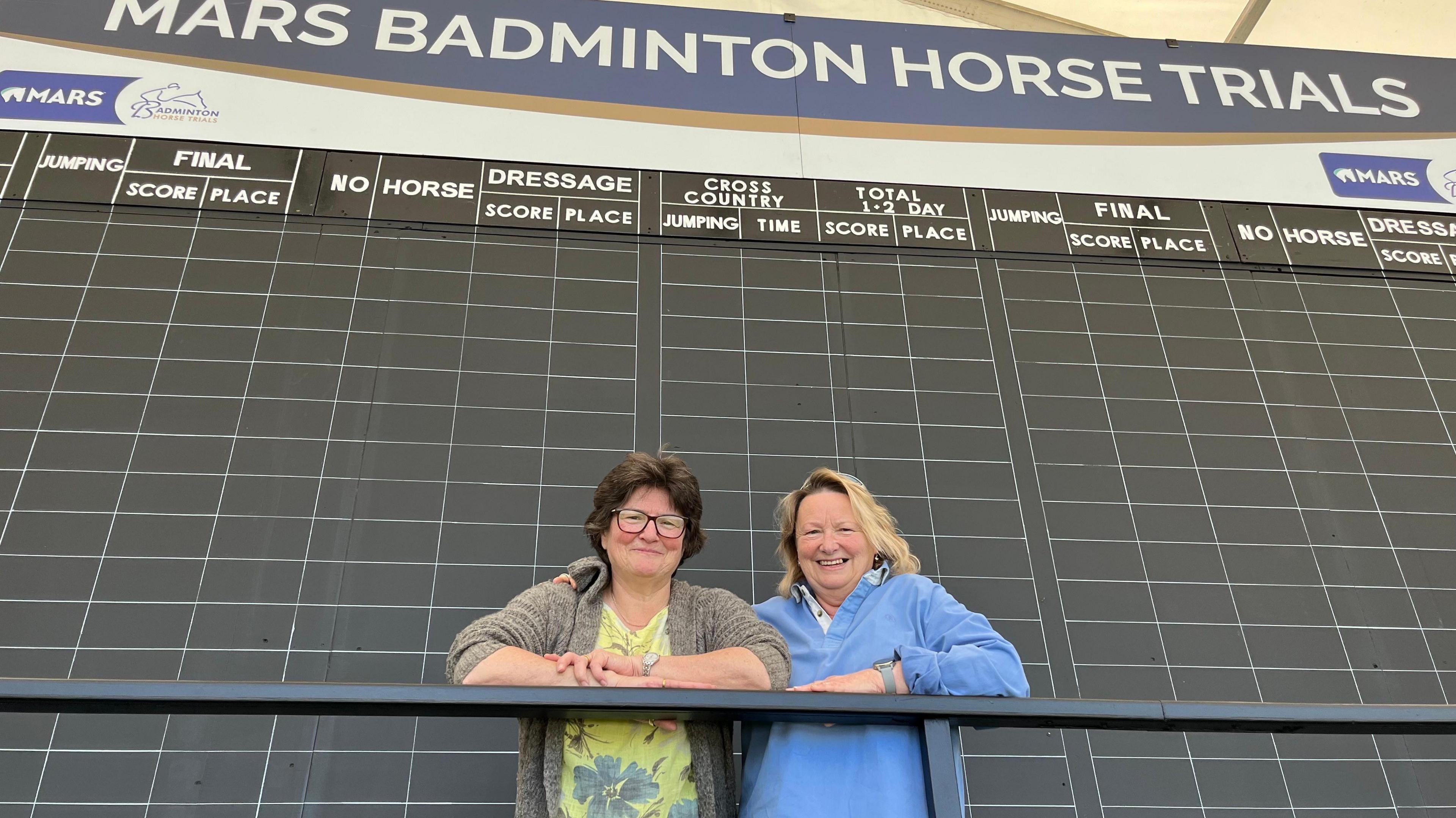 Sarah Tucker (r) and Badminton Horse Trials scoring assistant Wendy Barke say they wouldn't miss it for the world as they stand in front of a huge blackboard full of squares to be filled in with score numbers.