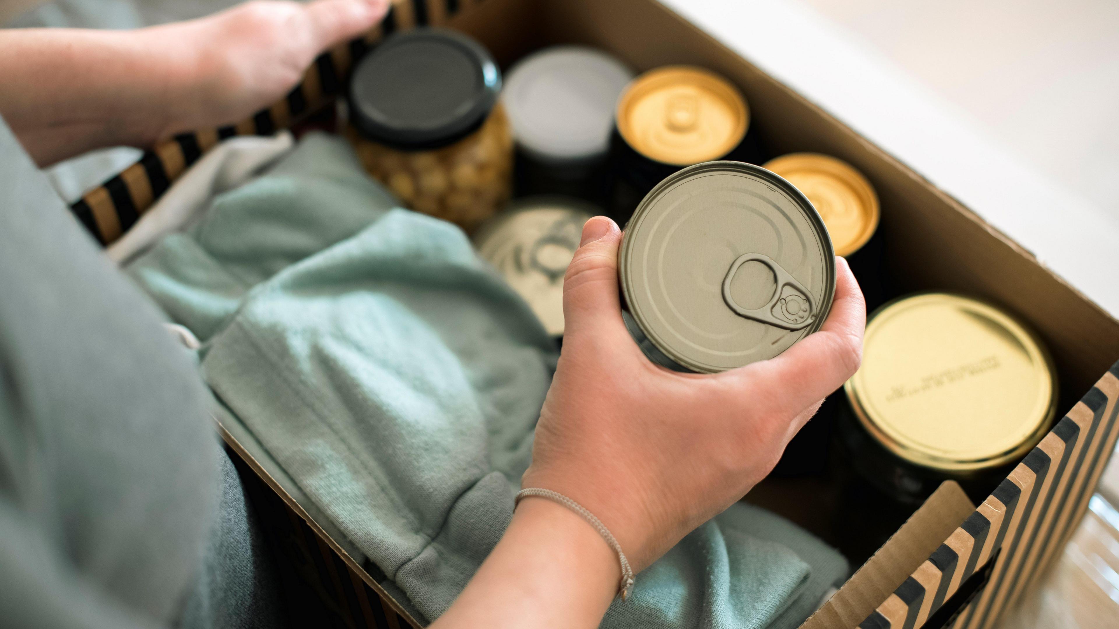 A woman's arms are in frame. She is holding a tin of food. Beside her is a cardboard box of food in tins and glass jars.