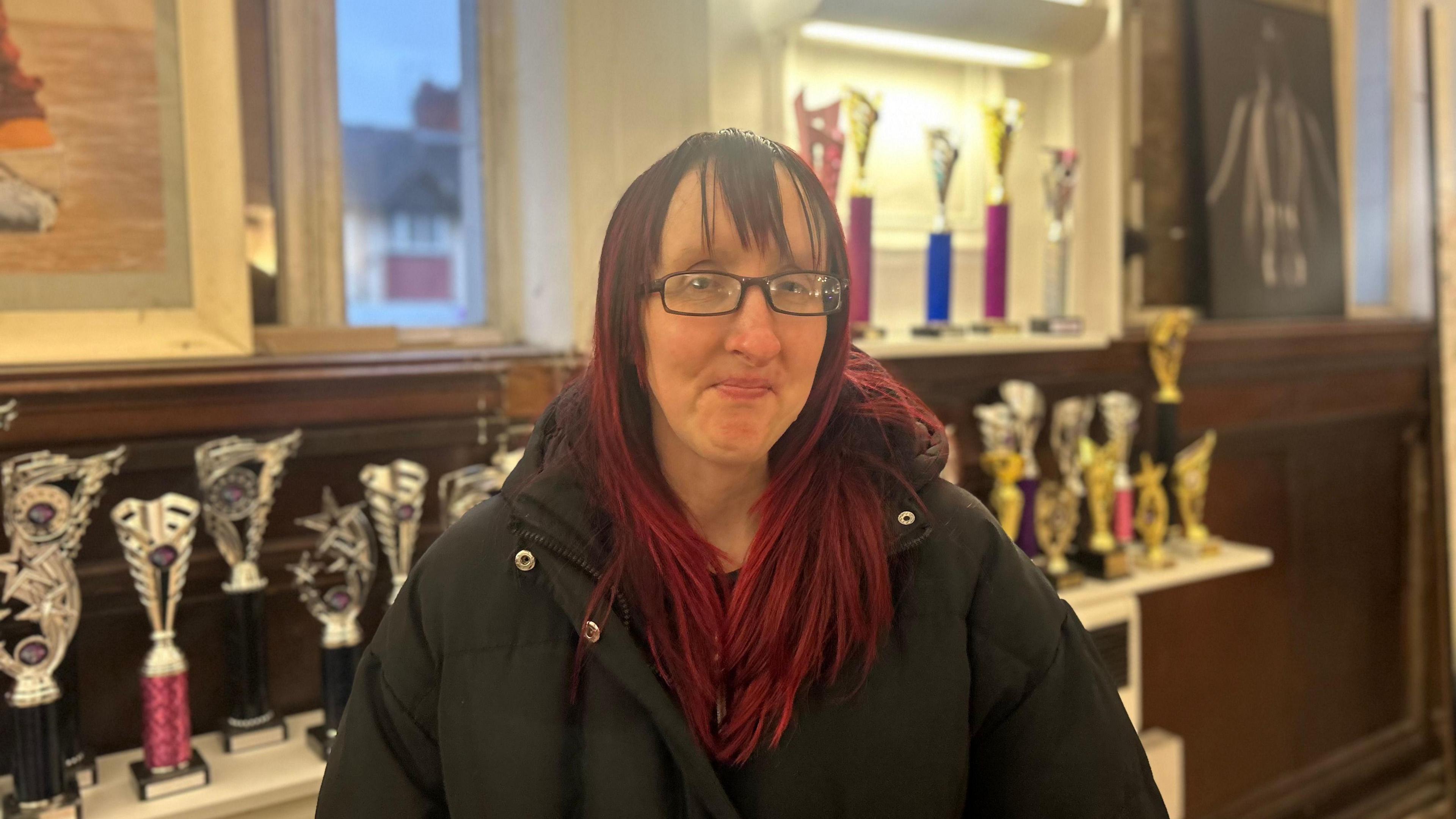 A woman with red hair and glasses standing inside the dance studio. She is wearing a black coat, and behind her are a number of shelves with dance trophies on them.